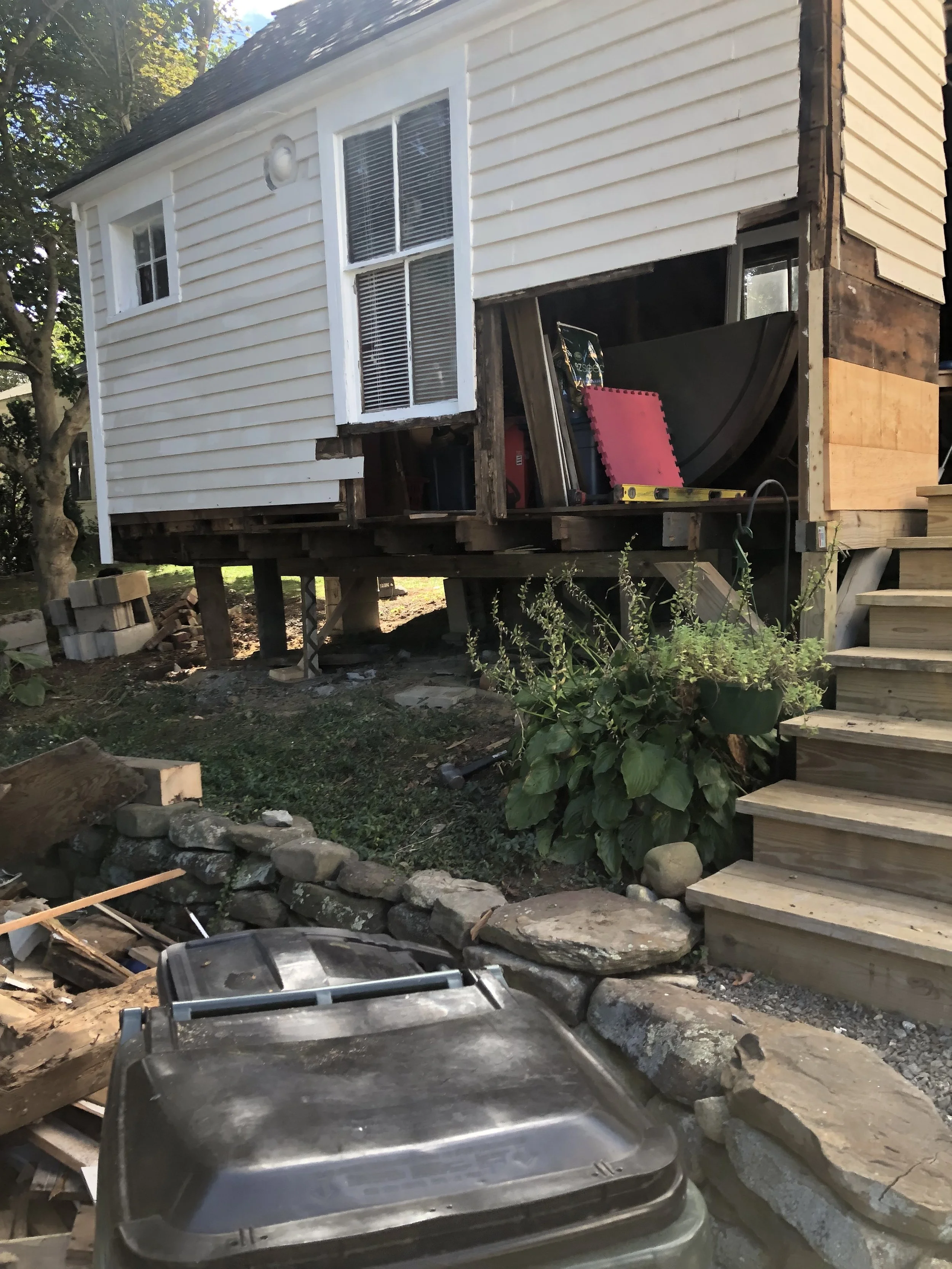 Front view of a house under renovation, with the porch partially removed and construction materials around. Wooden stairs lead up to the house with white siding, and a large plant is near the stairs. A trash bin is in the foreground.