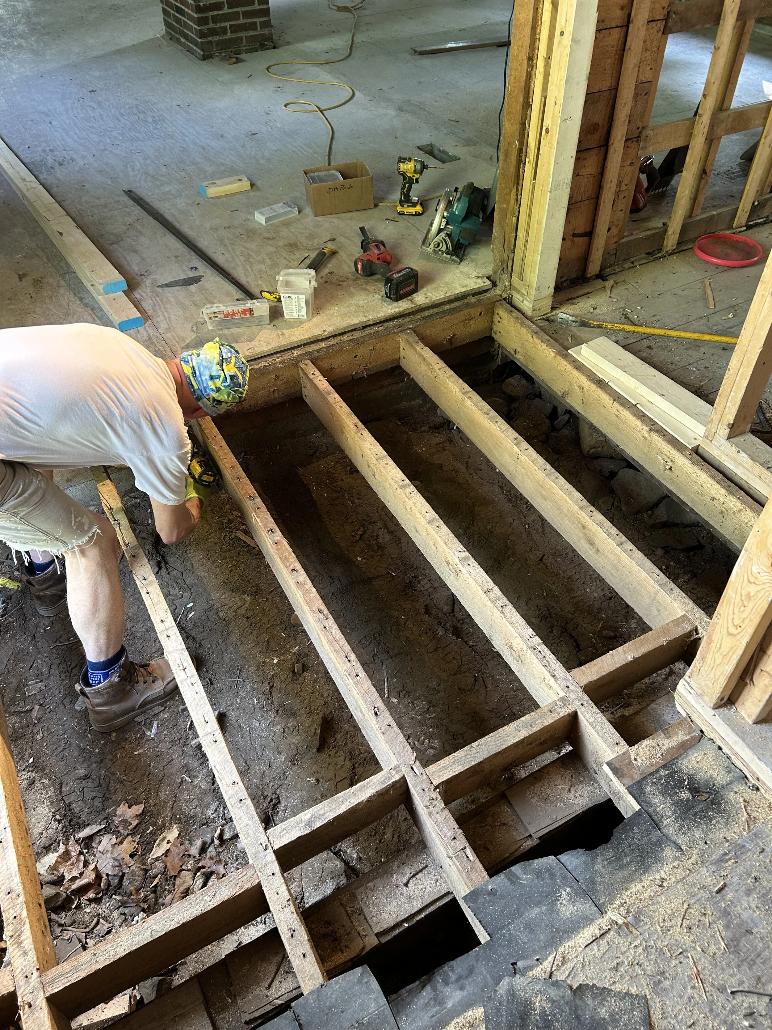 A construction worker installing a new floor in a house. The floor has been removed, exposing the wooden joists beneath. Various tools are scattered on the adjacent platform, including a drill, a saw, and measuring tape.