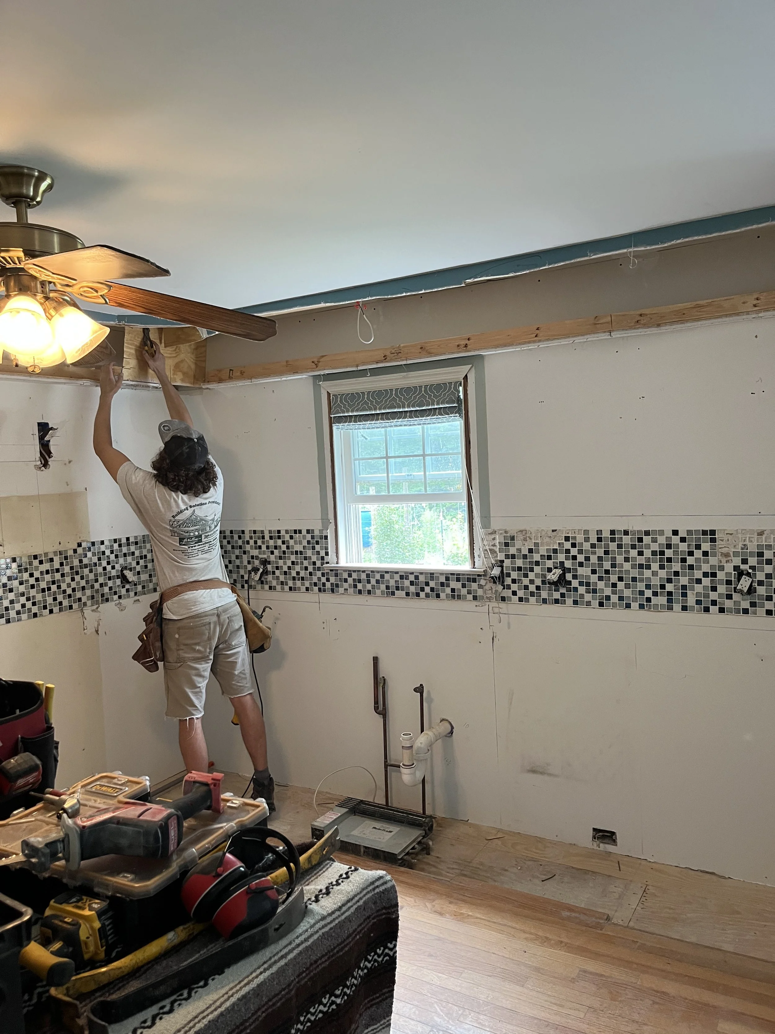 Person installing a wooden beam near the ceiling in a kitchen under renovation, with tools on a table in the foreground.