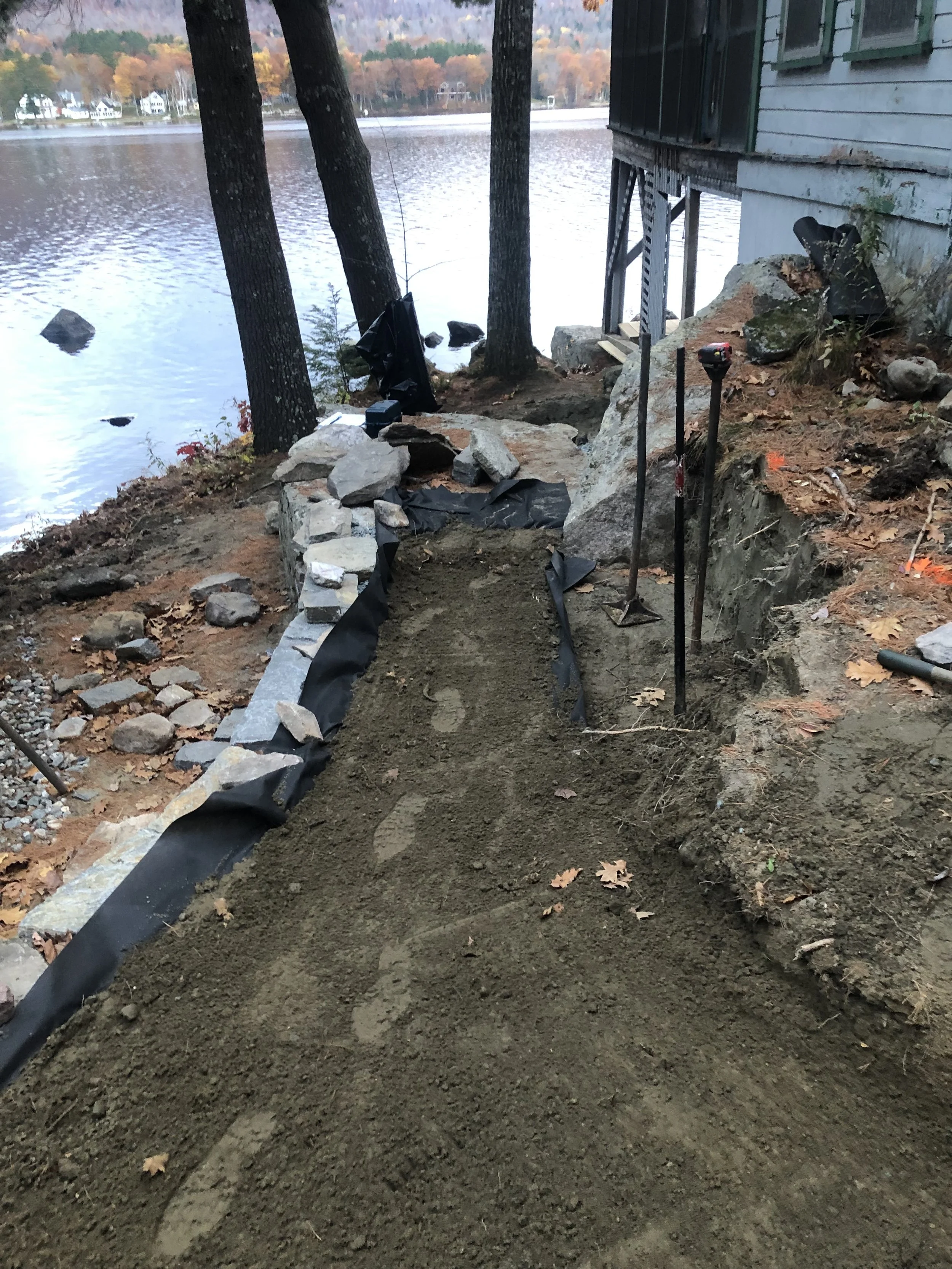 Construction site along a lakeside where a new pathway or border is being built with dirt and rocks, adjacent to a house with trees in the background.