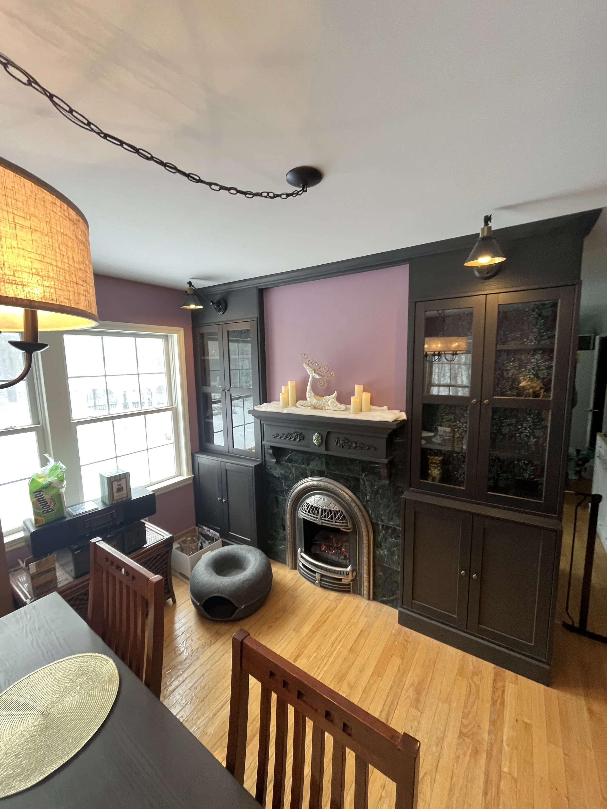 Living room with a black fireplace, dark wooden cabinets, pink wall, large window, and a gray cat bed on a wooden floor.