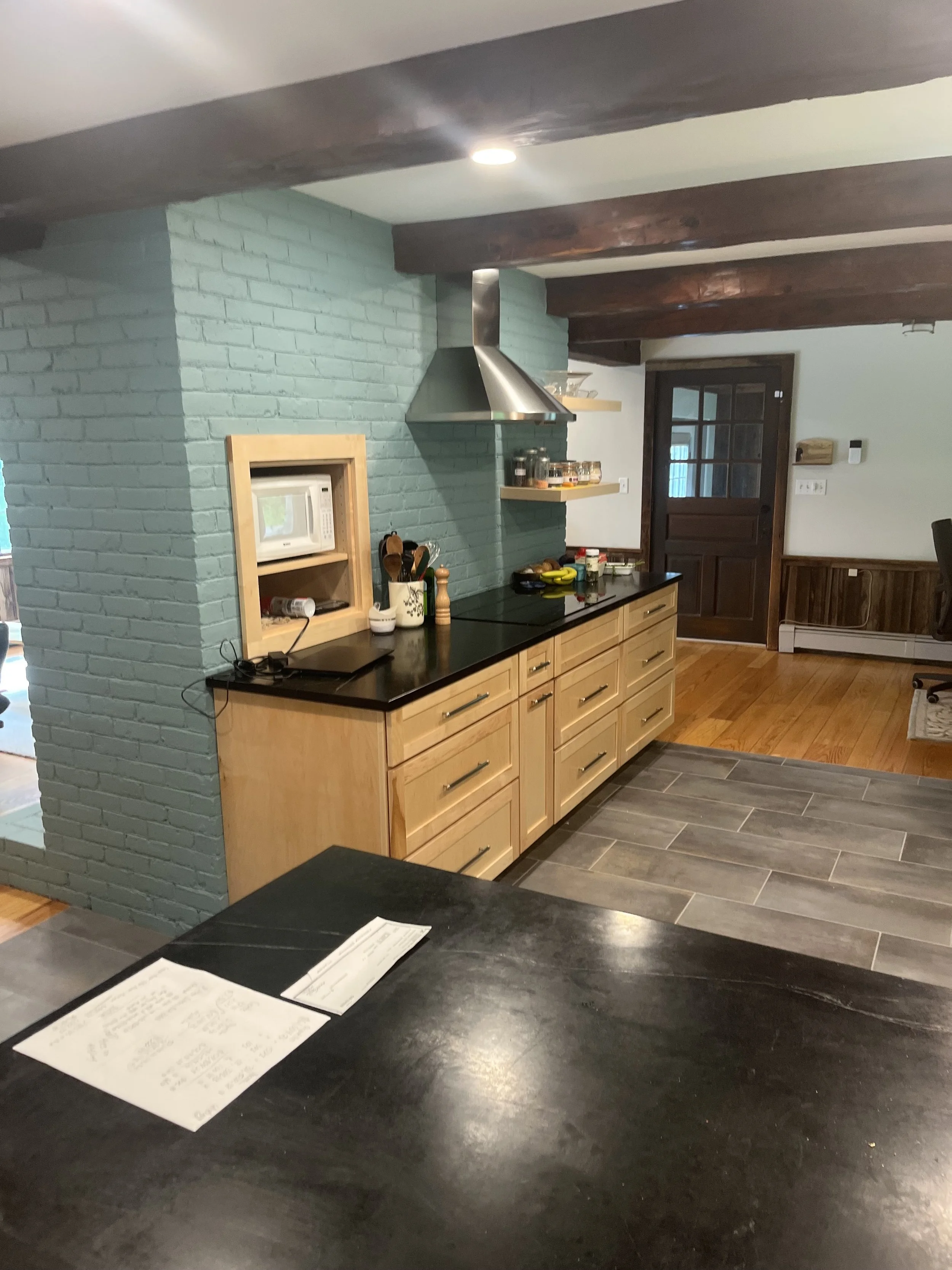 Kitchen with wooden cabinets, black countertop, stainless steel range hood, teal brick wall, and wood and tile flooring.
