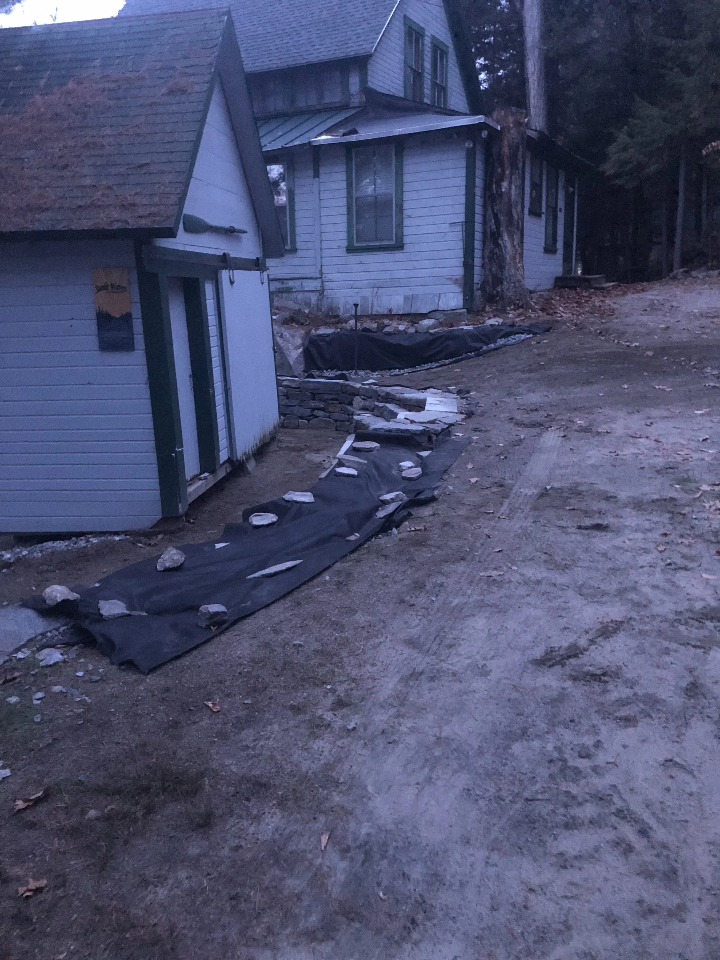 A rural house with a small outbuilding, both with weathered white siding and green trim. A pathway lined with rocks and black landscaping fabric leads to the house, with leaves scattered on the ground and trees in the background.