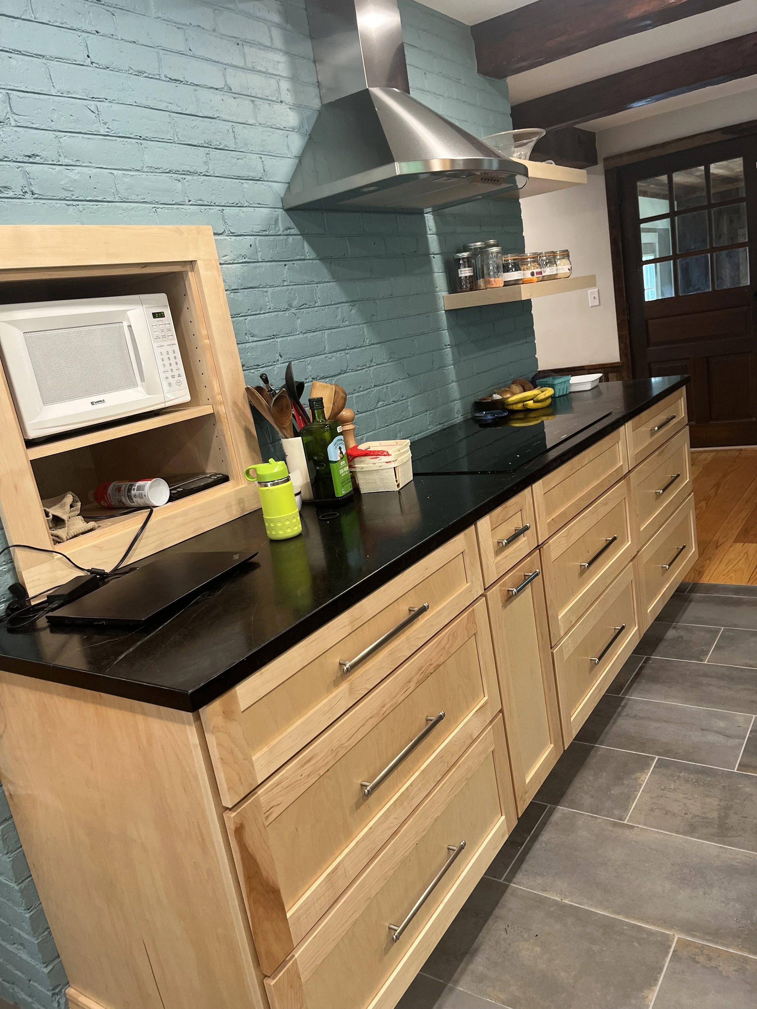 Kitchen with light wood cabinets, black countertop, teal brick wall, microwave on a shelf, stovetop, stainless steel range hood, and various small kitchen items and bananas on the counter.