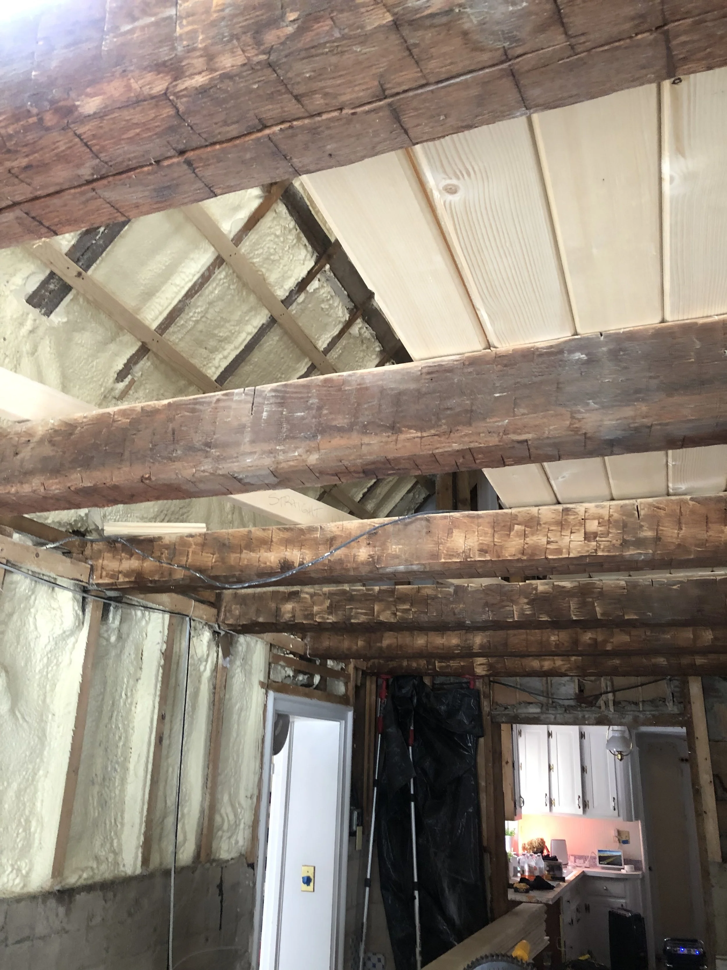 Interior view of a house under construction, showing exposed wooden beams, wall insulation, and partially finished ceiling with plywood panels.