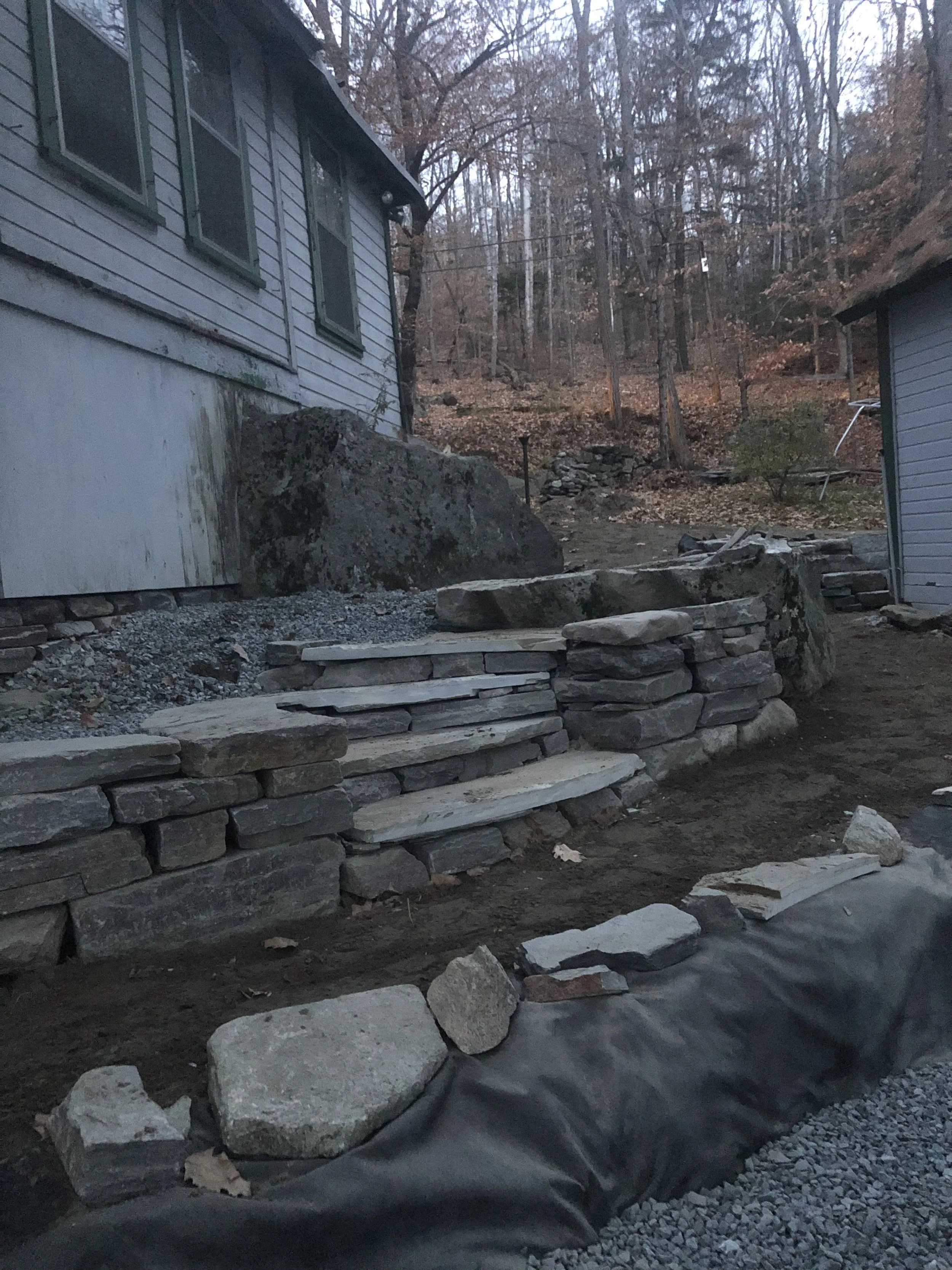 Stone steps and retaining wall under construction between two houses, with a wooded forest in the background.