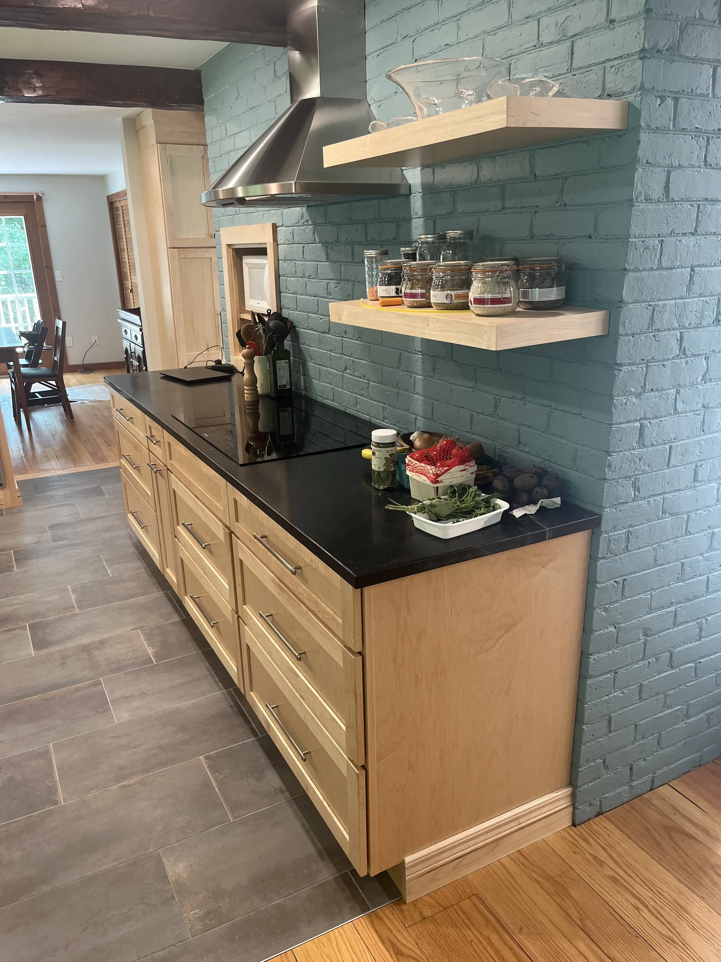 Kitchen with a row of wooden drawers, black countertop, teal brick wall, floating shelves with glassware and jars, and a stove with a stainless steel range hood.