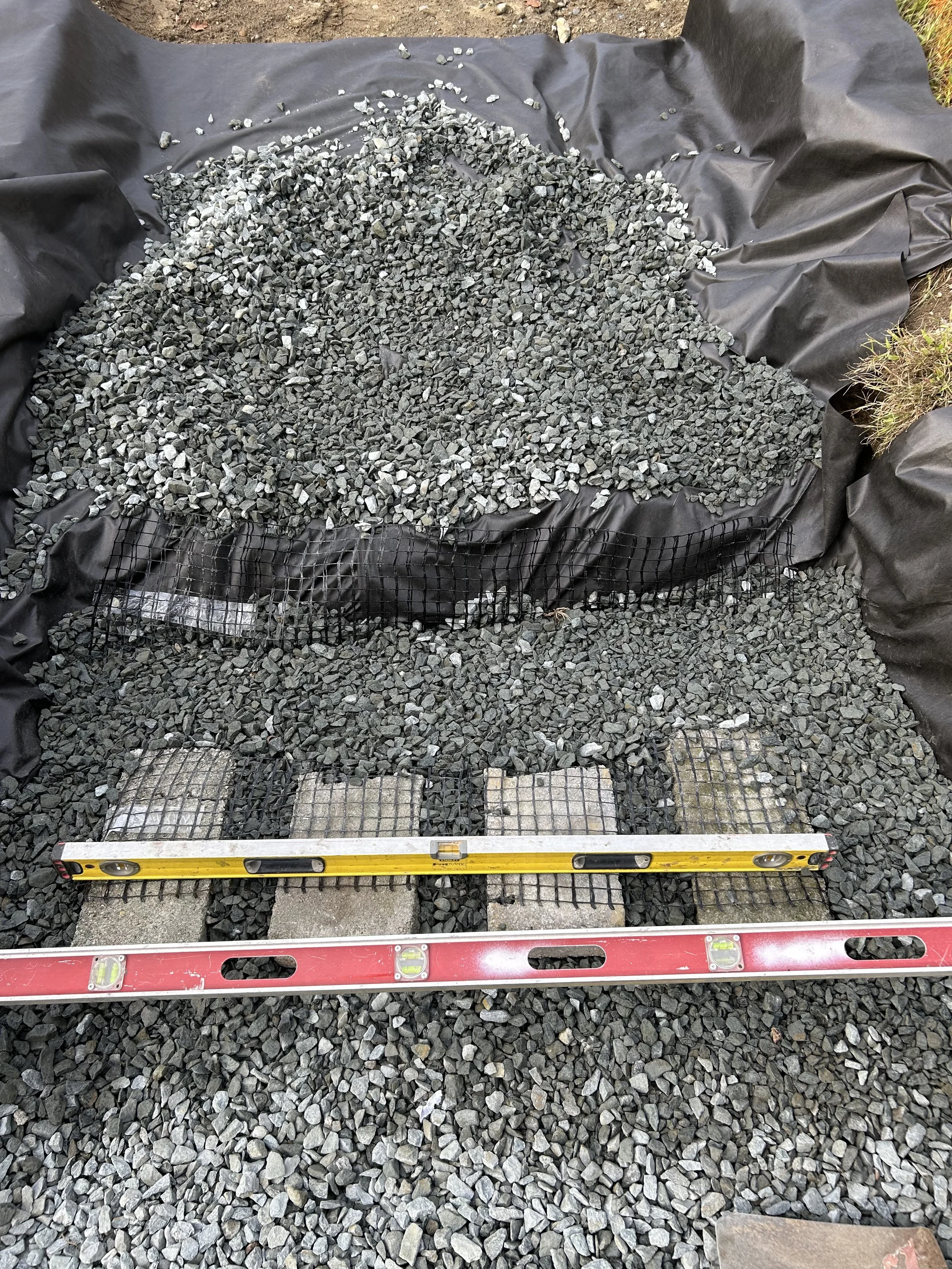 Landscape view of a construction site with black fabric, gravel, and concrete pavers, with a level tool on top.