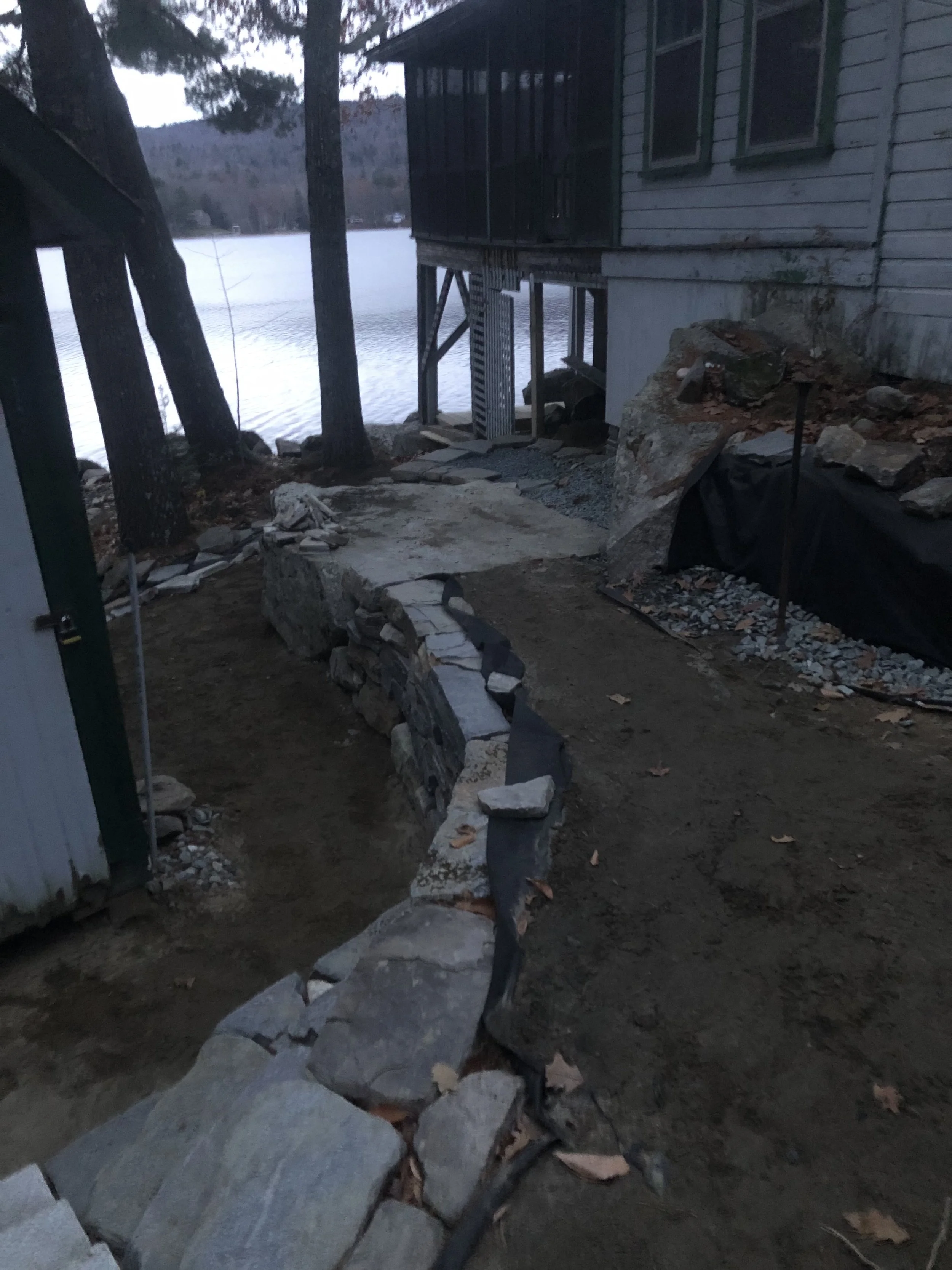 A construction site near a lakeshore shows a partially built stone retaining wall, surrounded by dirt and gravel. In the background, there are trees and a body of water, with a house elevated on stilts. The setting appears to be in a natural, outdoor