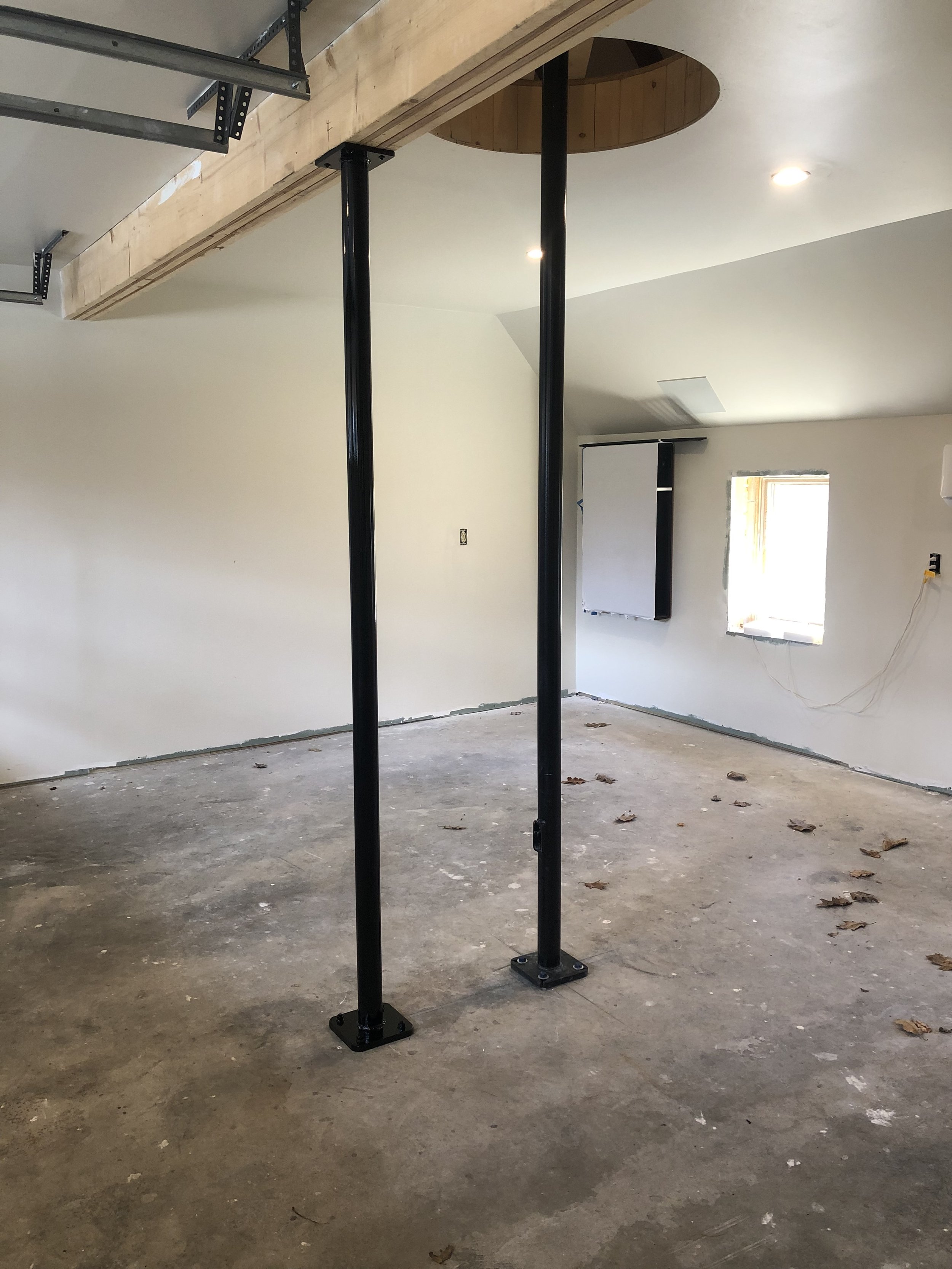 Interior room under construction with exposed concrete floor, white walls, small window, and two black metal support poles. There is a circular cut-out in the ceiling with unfinished wood, a partially installed wall mounted screen, and some wiring on