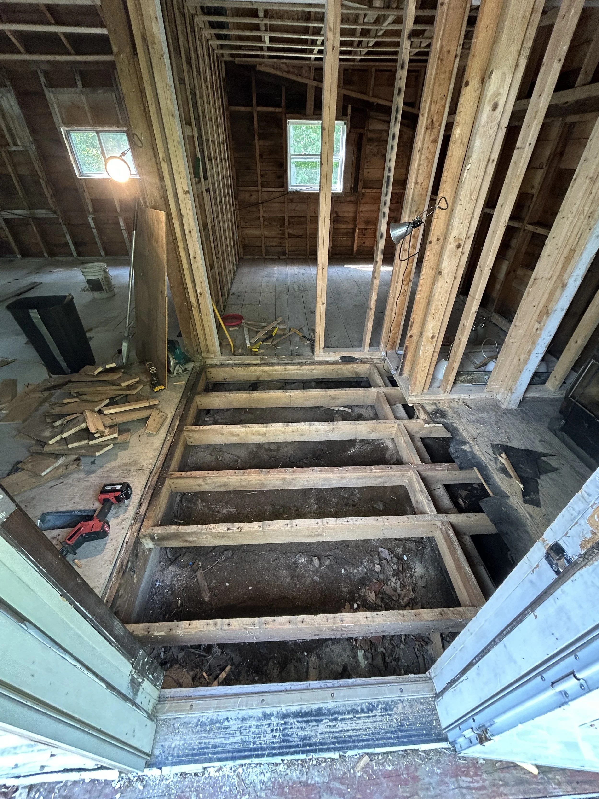 Interior view of a home under renovation, showing exposed wooden floor joists and framing for a new floor or wall, with construction tools and materials scattered around.