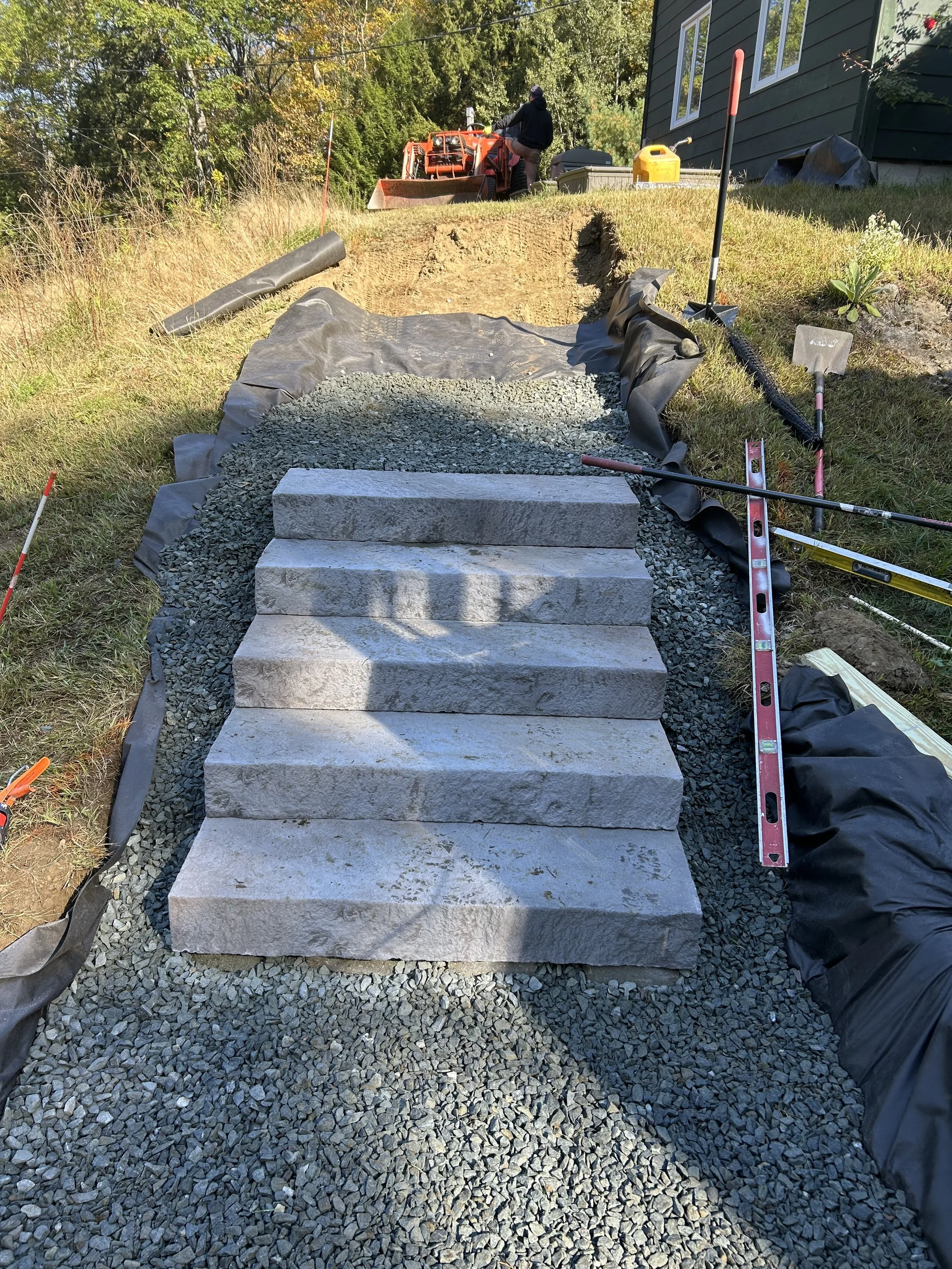 Construction of a stone staircase outdoors with tools and materials around, a worker operating machinery in the background.