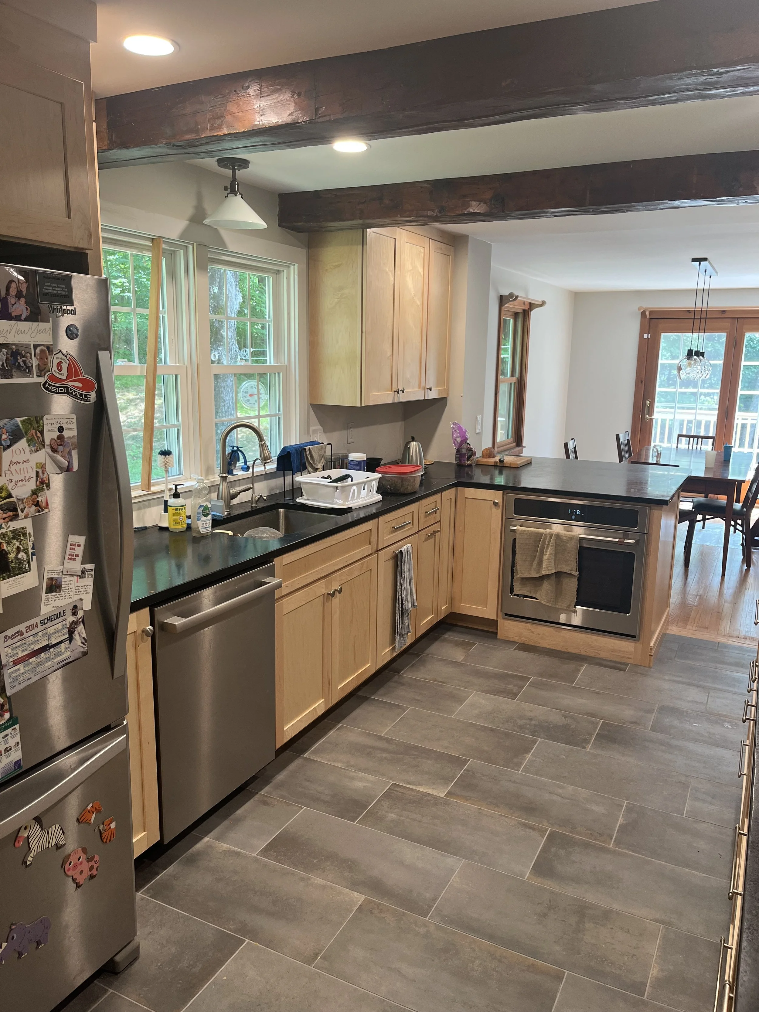 Kitchen with wooden cabinets, black countertops, stainless steel appliances, tiled floor, and a window with a view of trees outside. There is a refrigerator with magnets, a sink with dishes, and a dining area with a table and chairs in the background