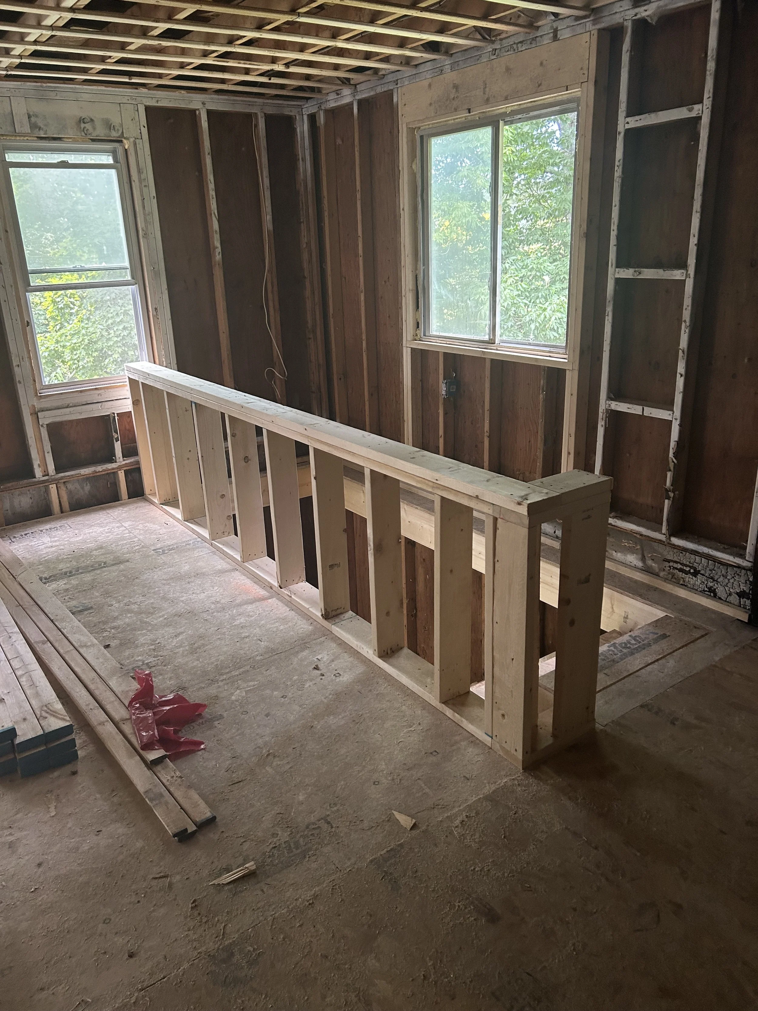 Interior of a house under construction, showing a staircase with new wood framing, two windows, and unfinished walls and ceiling.
