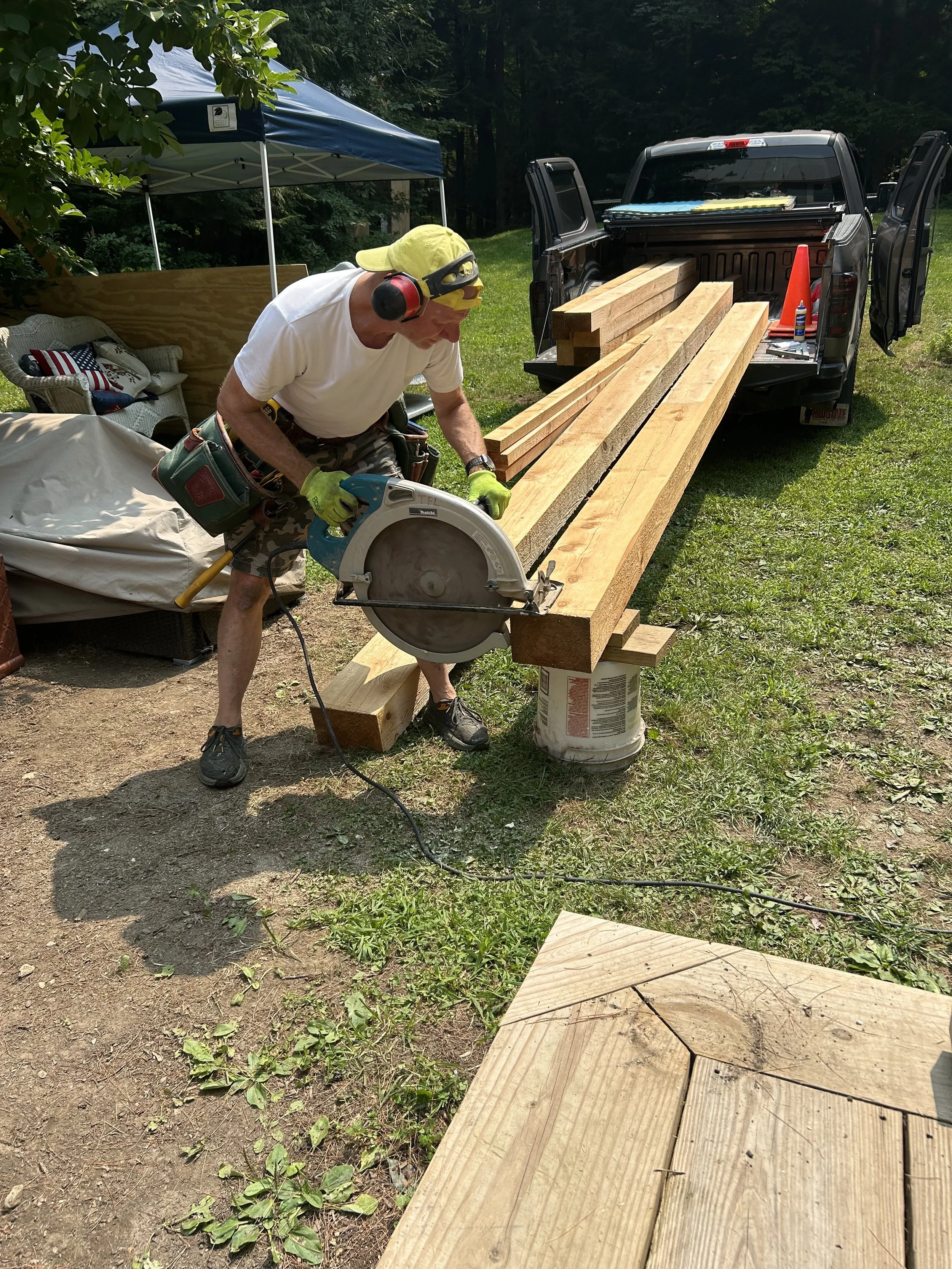 A man using a circular saw to cut a wooden beam outdoors, with a pickup truck and a canopy tent in the background.