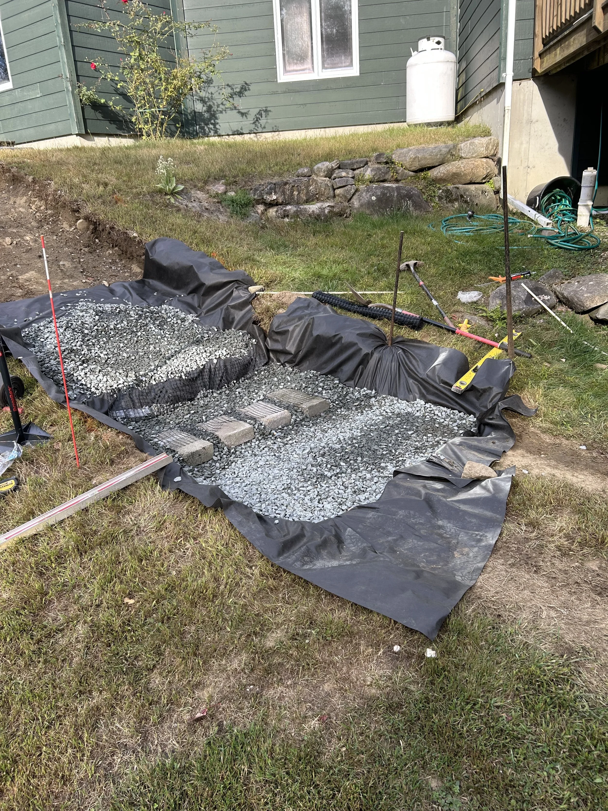 A small backyard area with a gardening project in progress. The project involves layering gravel and bricks on black landscape fabric, likely for a pond or garden feature. Tools such as a level, a hammer, and other gardening equipment are scattered a