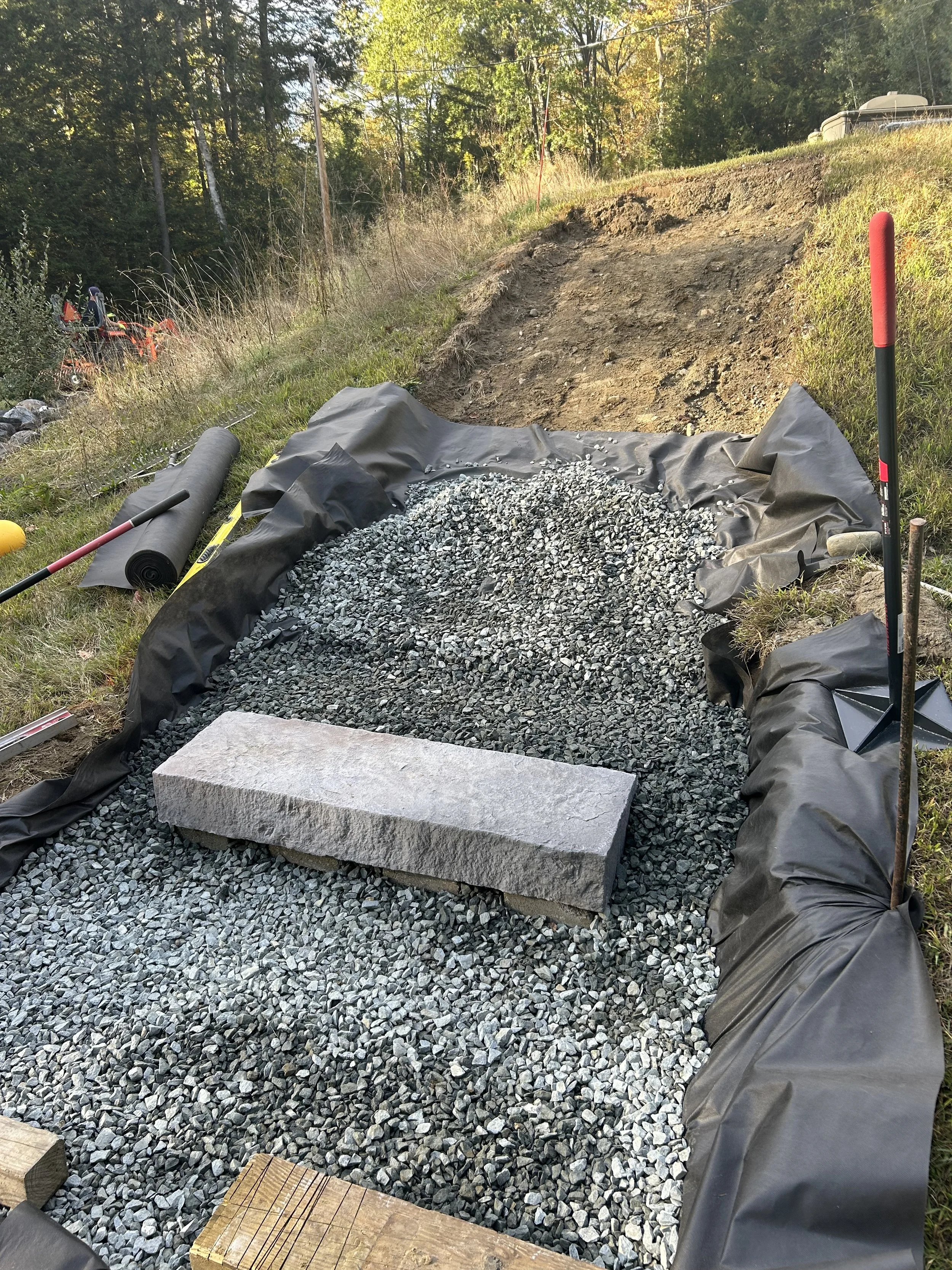 A construction site showing a gravel pathway lined with black geotextile fabric, with a concrete block placed on top, and construction tools nearby. The surrounding area is grassy with trees and an incline in the background.
