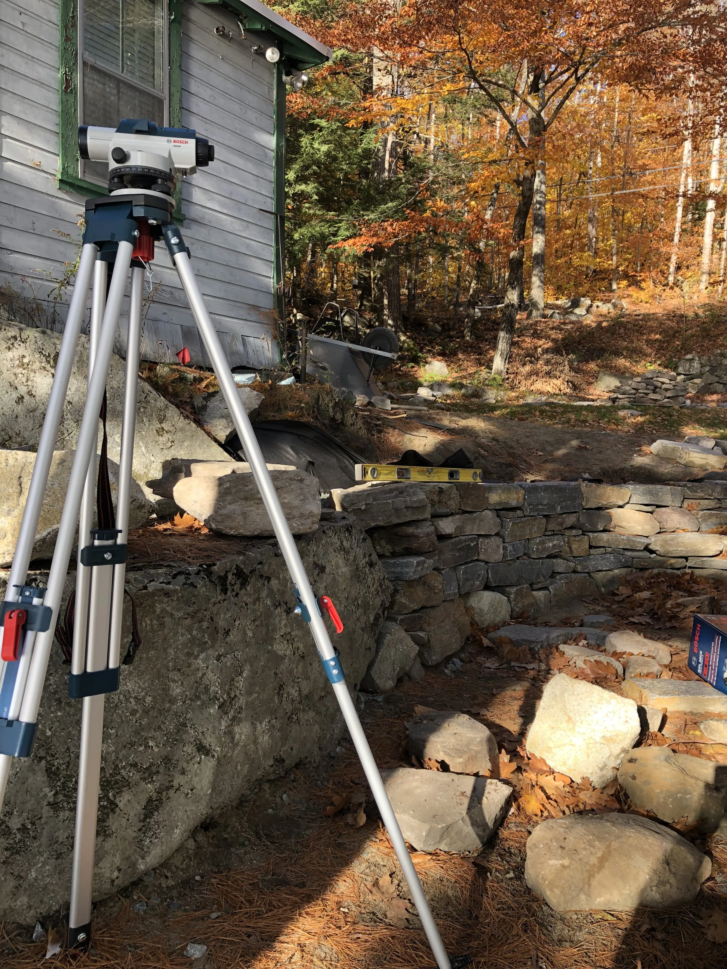 A surveying tripod set up outdoors near a stone wall with a level tool on top, with fall foliage trees in the background.