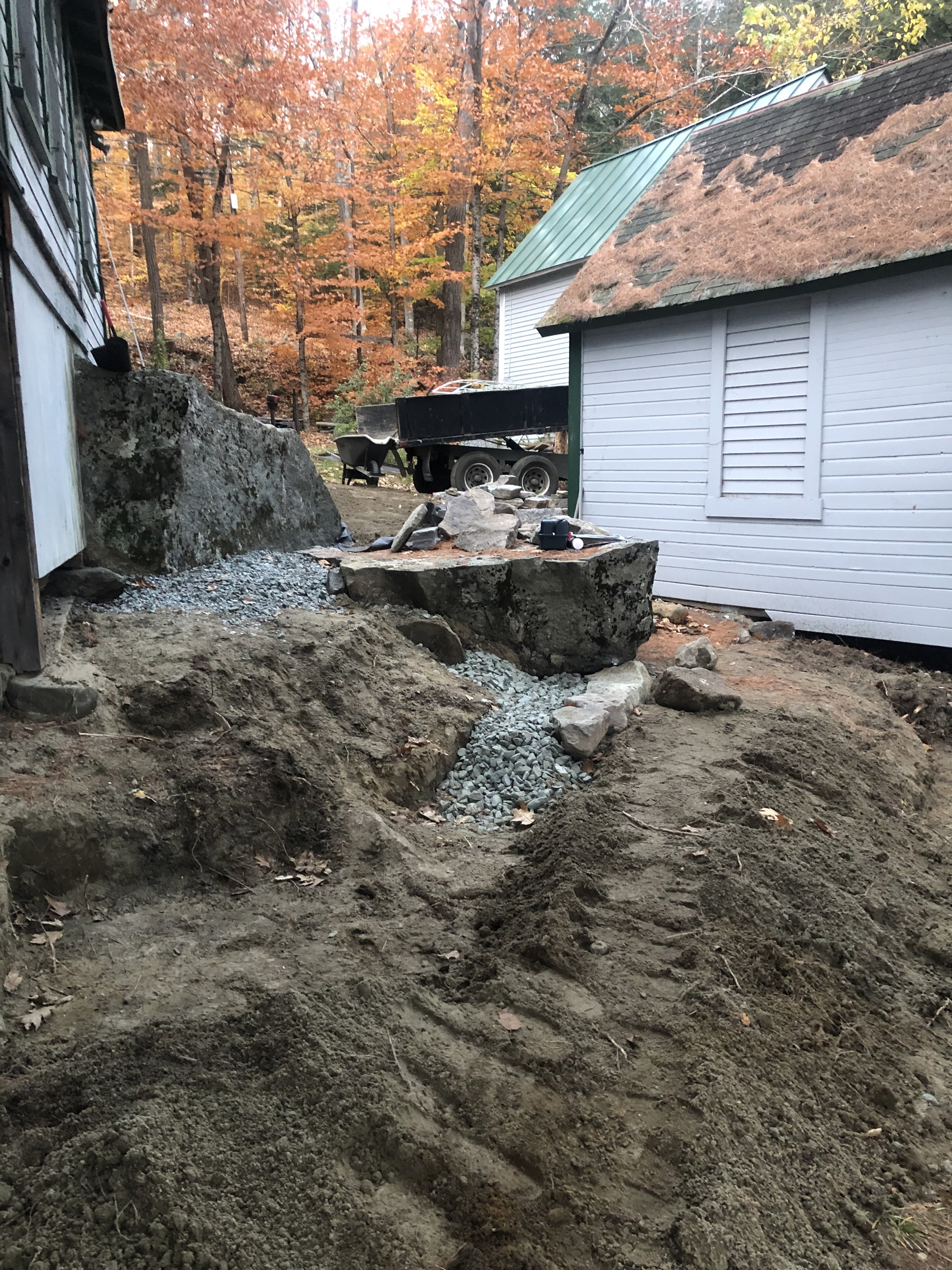 A construction site with dirt and gravel, large rocks, and a partially dug trench next to a house with trees with orange and yellow autumn leaves in the background.