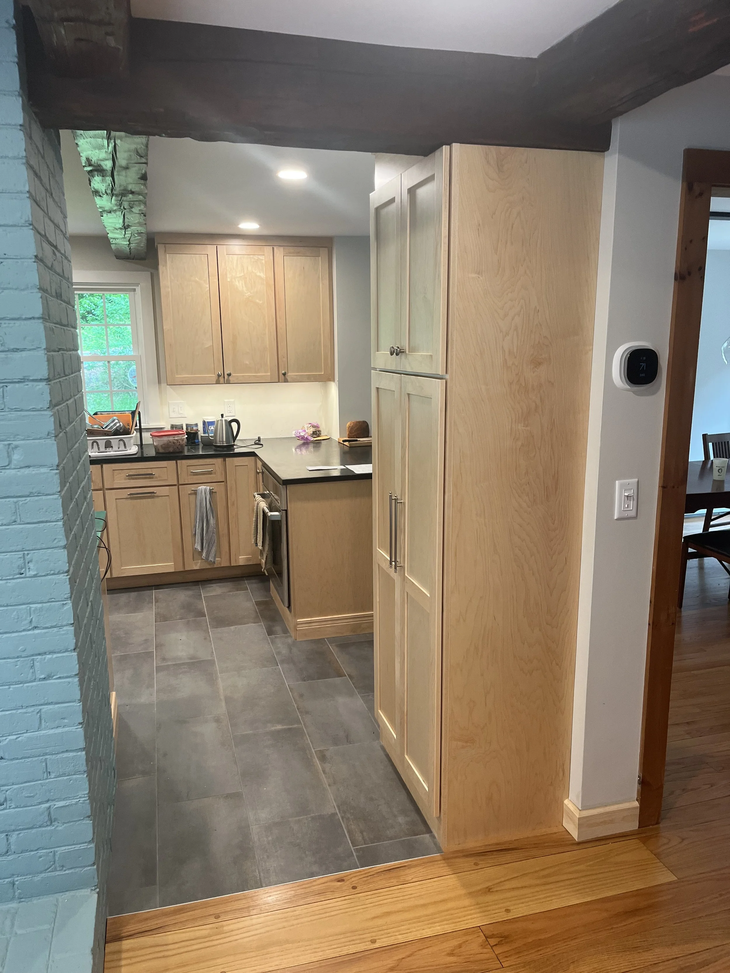 View of a kitchen with wooden cabinets, a window with a green view, a black countertop, and gray floor tiles. Various kitchen items are on the counter, including a kettle, dishes, and a cutting board. A wooden doorframe is visible to the right.