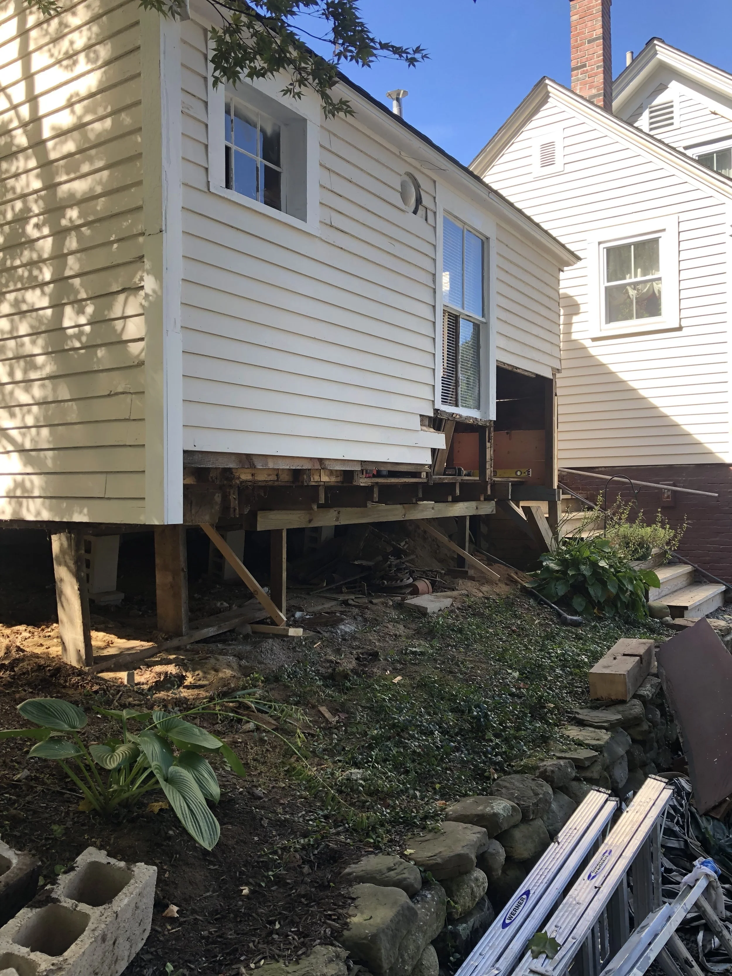 Side of a house under renovation with the foundation exposed, a set of stairs, and construction tools nearby.