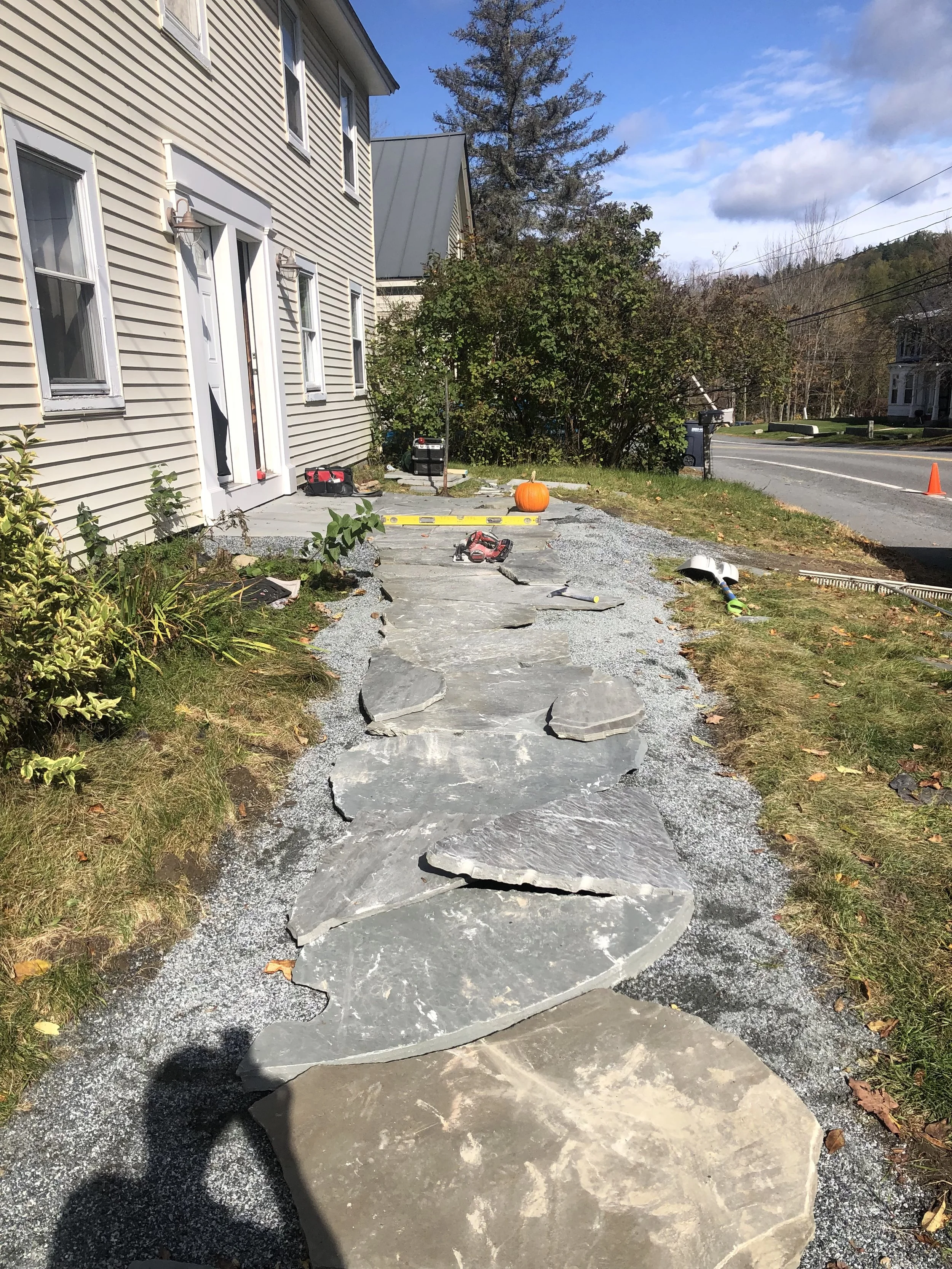 A sidewalk being built with large stone slabs in front of a house, with tools and a pumpkin visible, under a partly cloudy sky. Hardscaping.