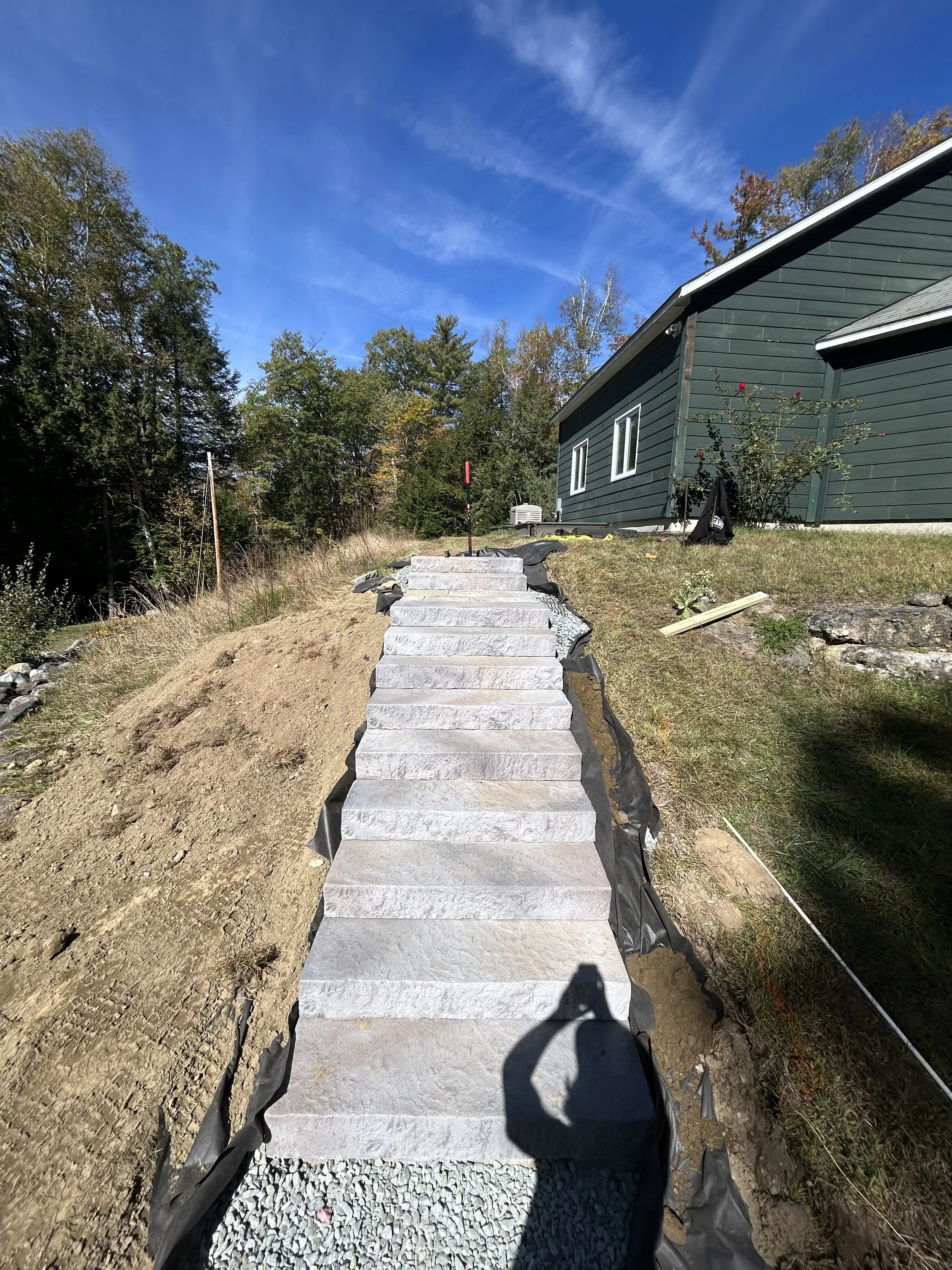 Stone staircase under construction on a hillside leading up to a house with green siding, trees in the background, and a partly cloudy blue sky.