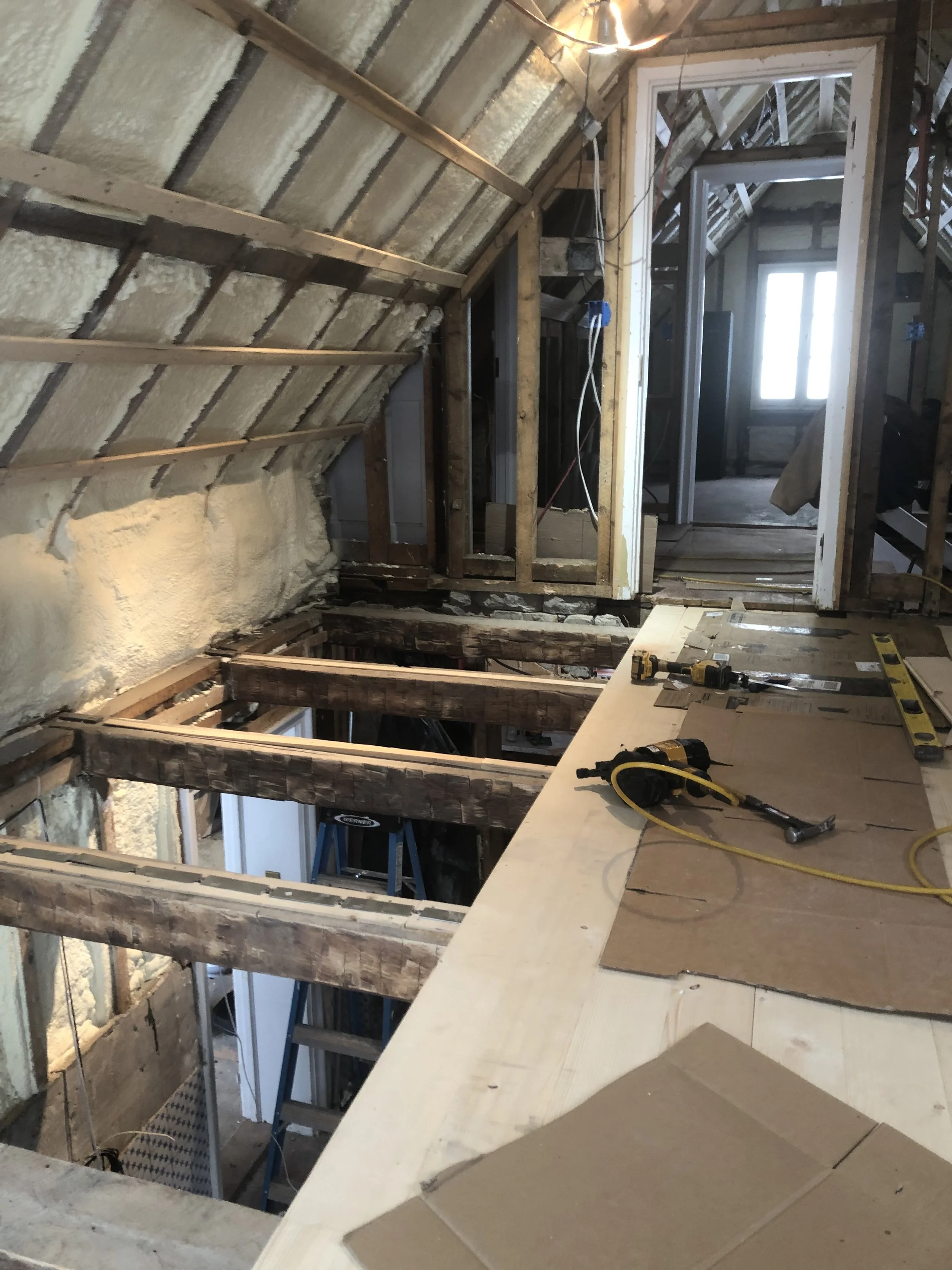 Attic under renovation with exposed wooden beams, insulation, construction tools on a makeshift work surface, and open door leading to a room with a window.
