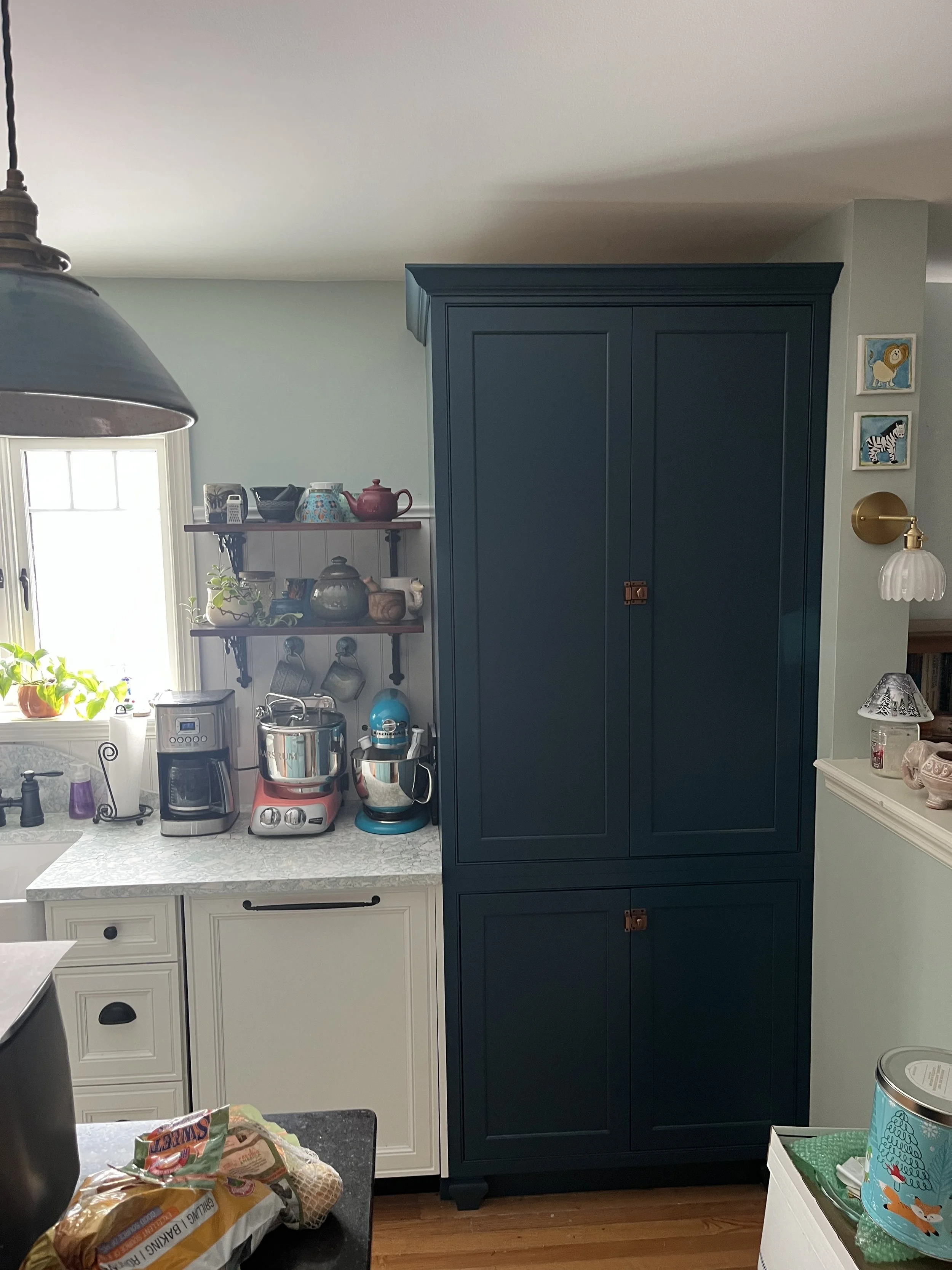 Kitchen with a large blue cabinet, a window with sunlight, two shelves with teapots, cups, and plants, and small appliances including a coffee maker, blender, and mixer on the counter.