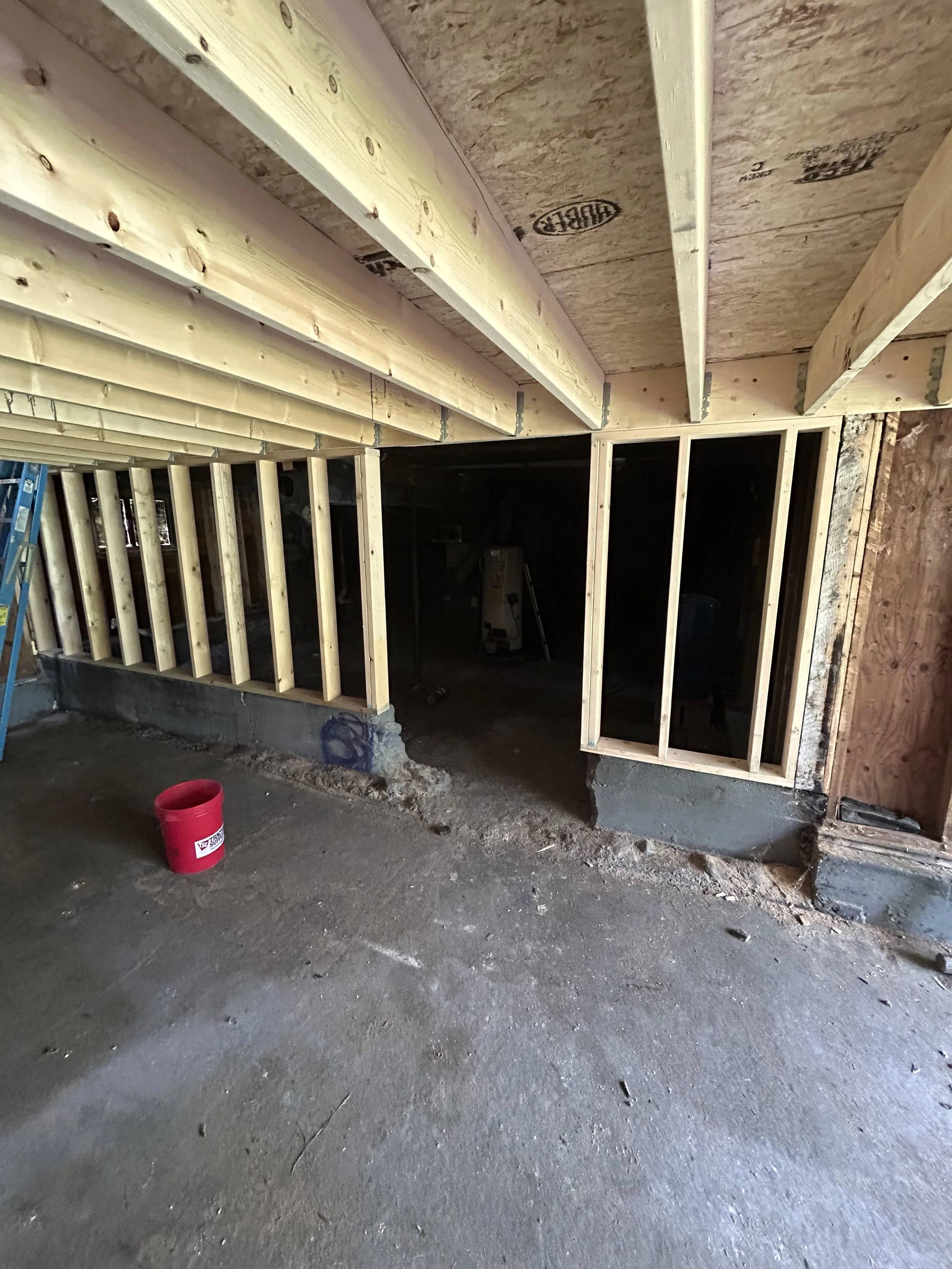 Construction site showing wooden framing in a building under construction, with a ladder on the left and a red bucket on the floor.