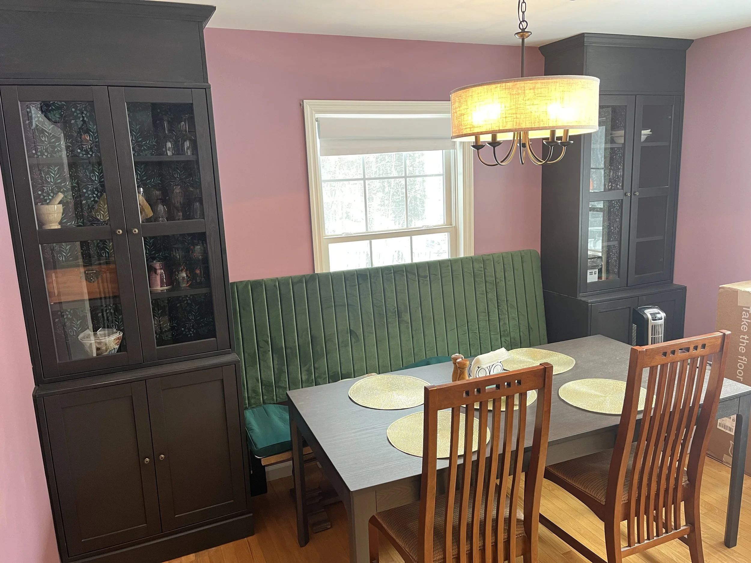 Dining room with pink walls, black china cabinets, green upholstered bench seating, wooden chairs, a window, and a chandelier.