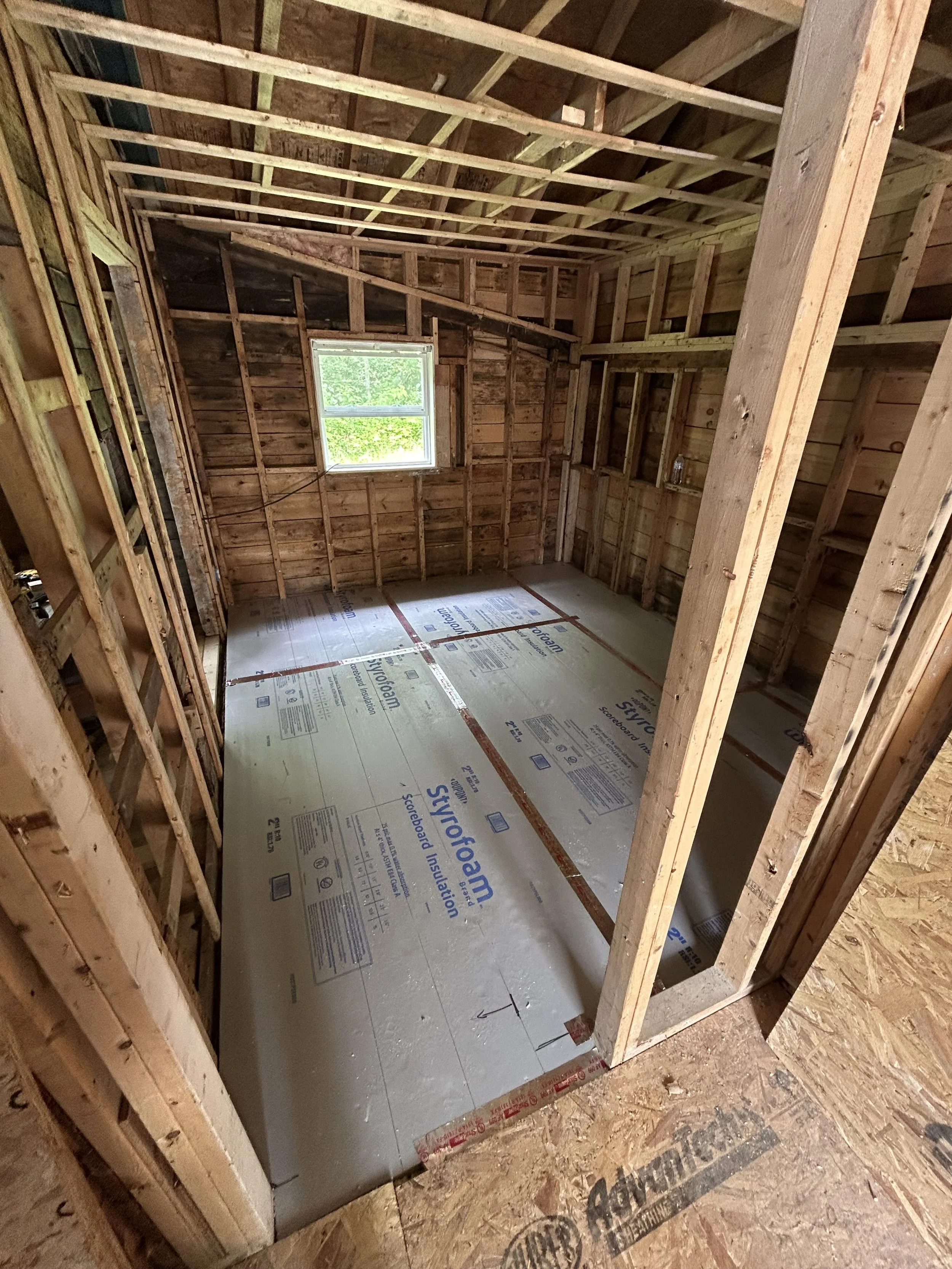 Construction of a room in a house with exposed wall and ceiling framing, a single window letting in natural light, and a subfloor with insulation panels ready for flooring installation.