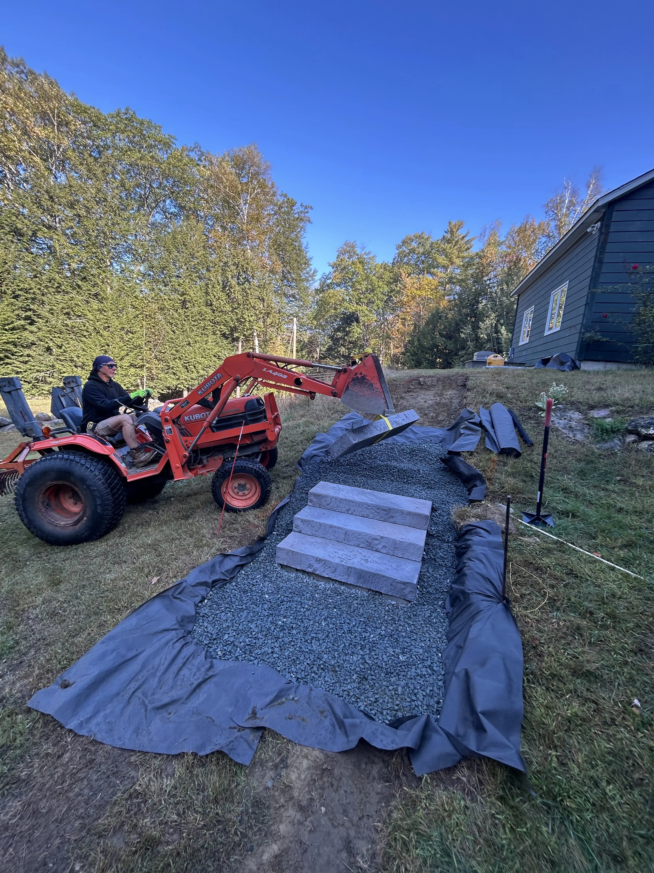 Person operating a red tractor with a front loader, moving gravel and stones into a garden bed or pathway on a sloped yard. Blue sky and trees in the background, neighboring house visible to the right.