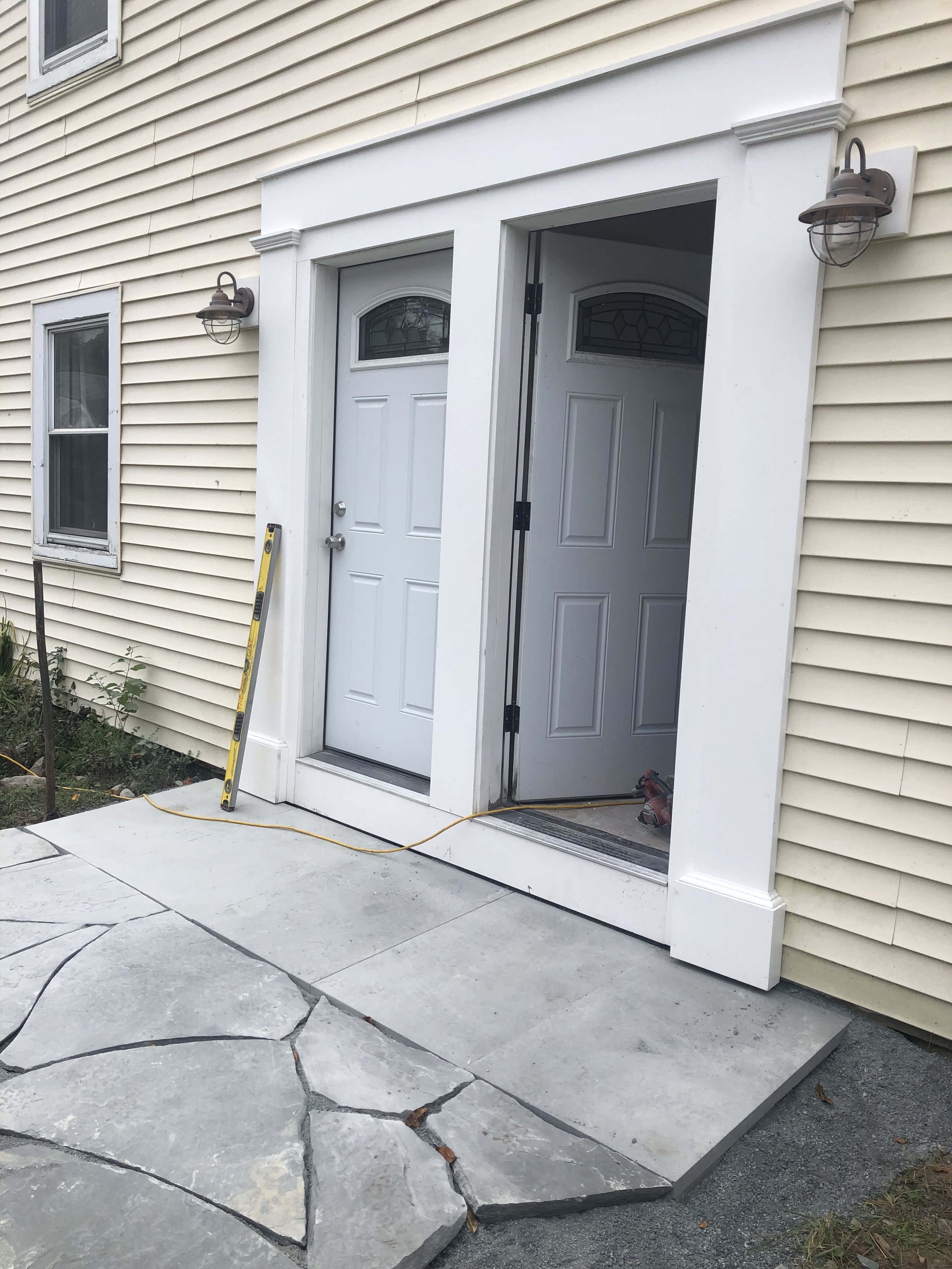 Doorway with two doors, one side open, with exterior lights on the wall and a level leaning against near the door, a yellow level and a yellow extension cord on the ground. Hardscaping.