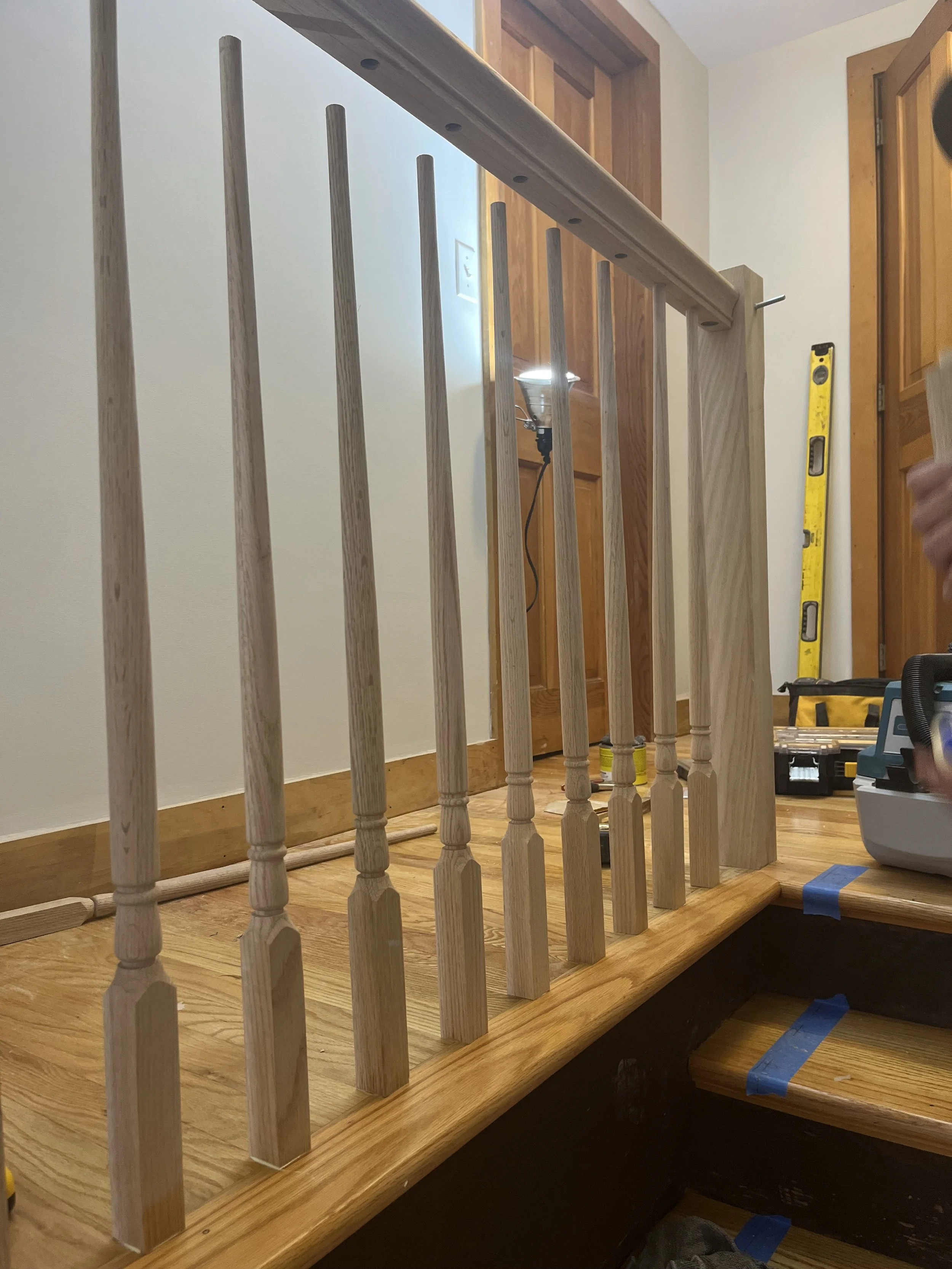 Close-up of a wooden staircase railing under construction with vertical spindles, a wooden handrail, and construction tools visible in the background.