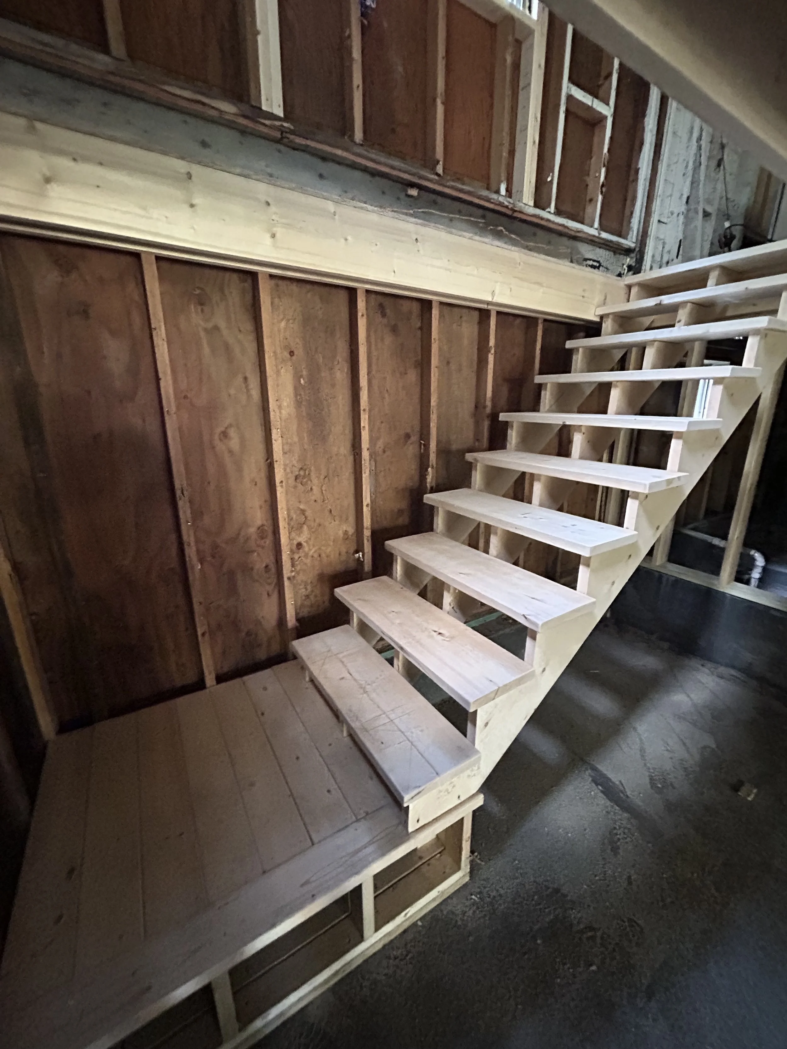 Wooden staircase with open risers inside an unfinished room with exposed wooden wall studs and framing.