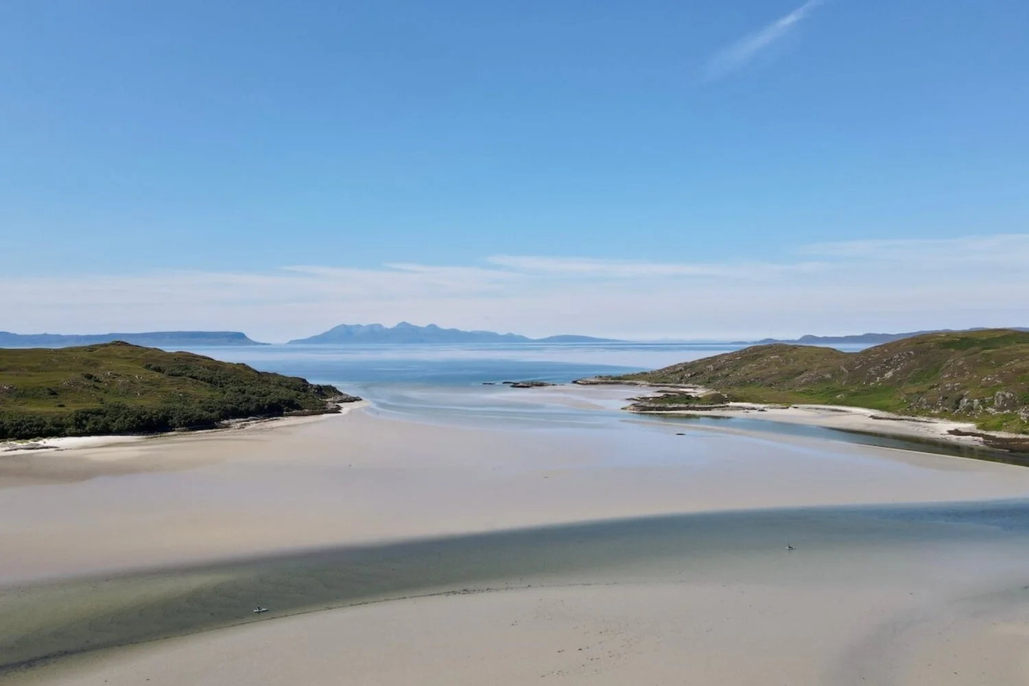 A panoramic aerial view of the silver sands of Arisaig beach at low tide, looking out across calm blue coastal waters toward the distant peaks of Eigg and Muck under a clear summer sky.