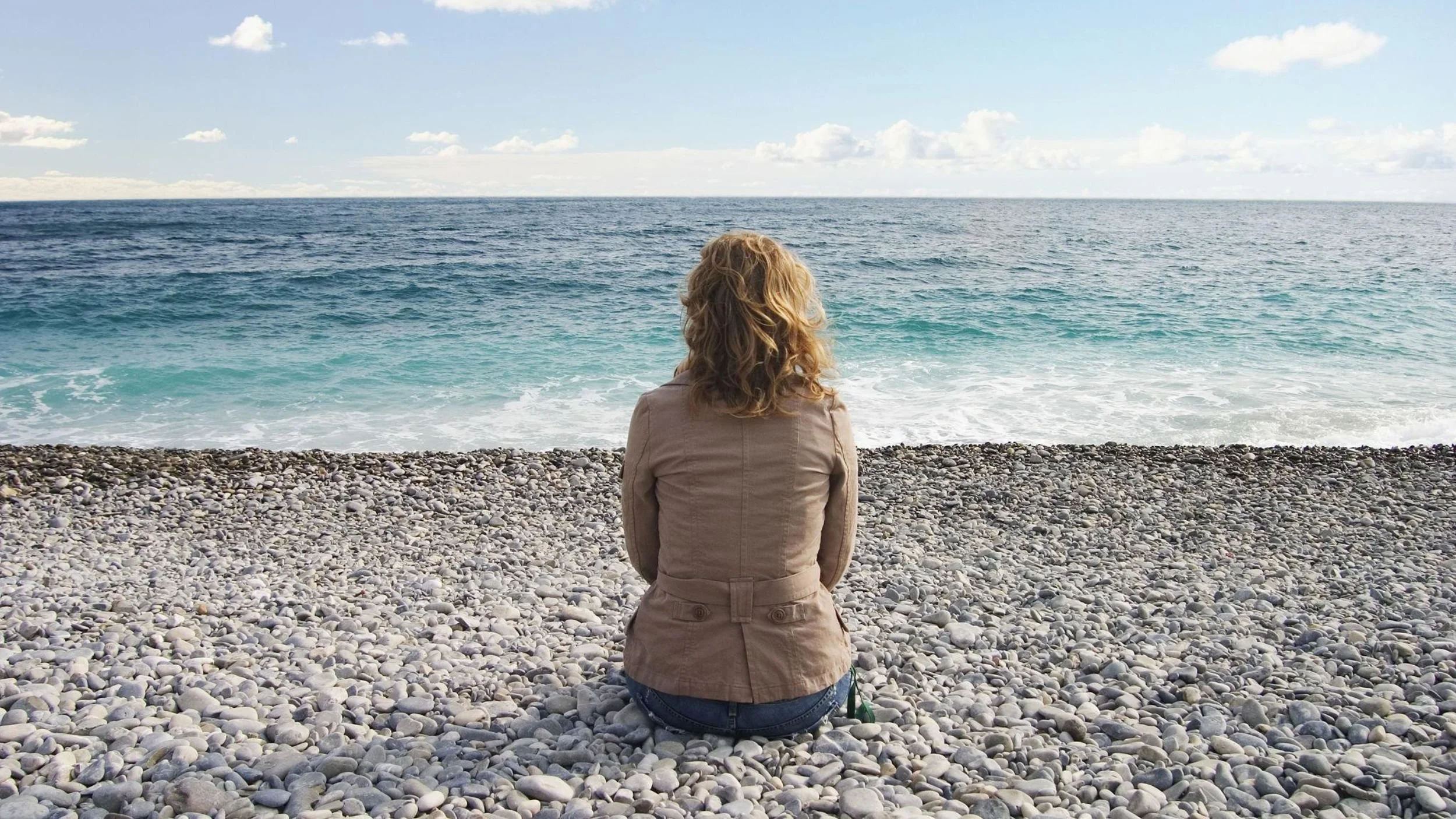 Woman sitting on a pebble beach looking out across the sea.