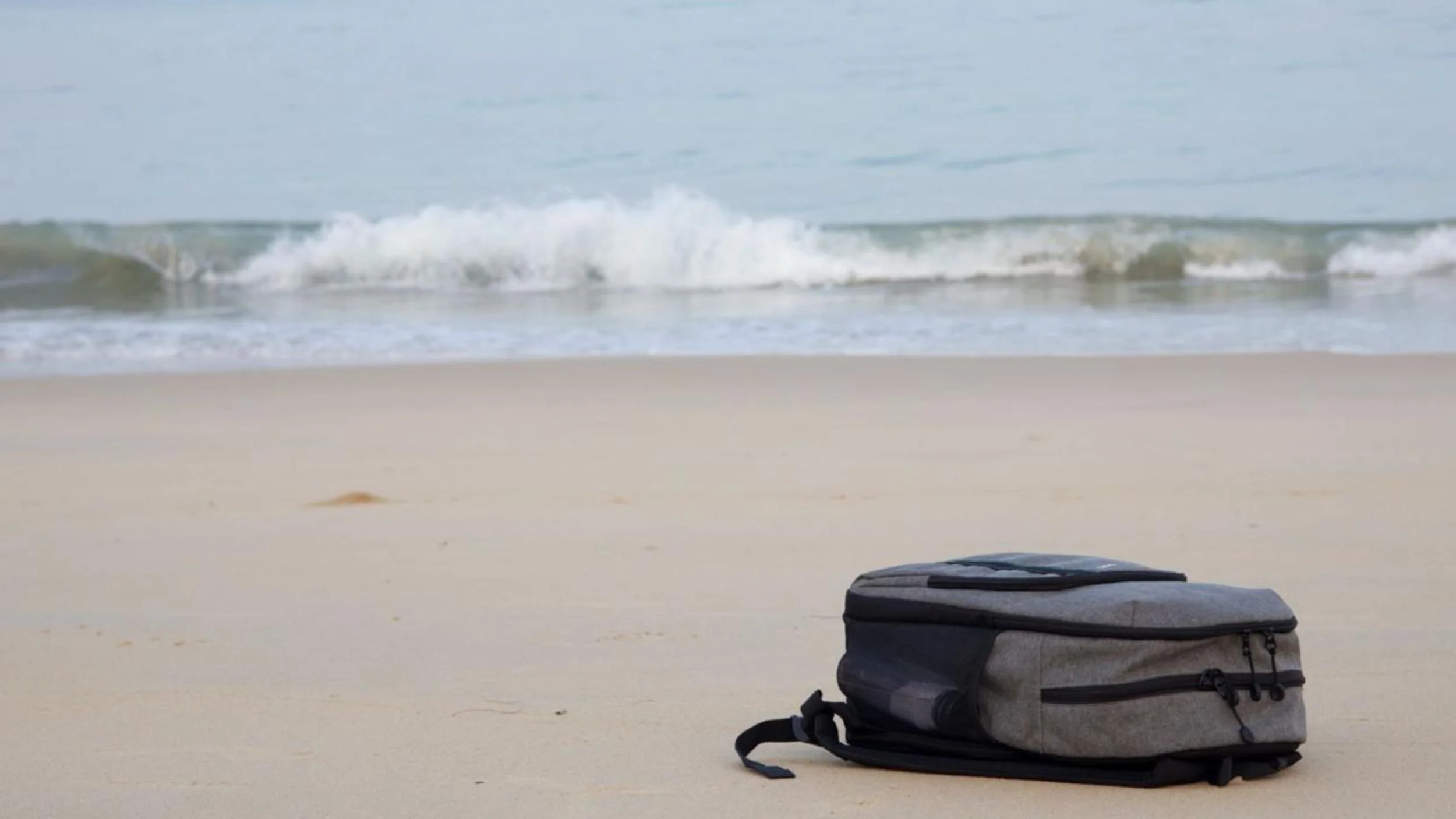 Grey bag resting on the sand beside the sea as waves break in the background.