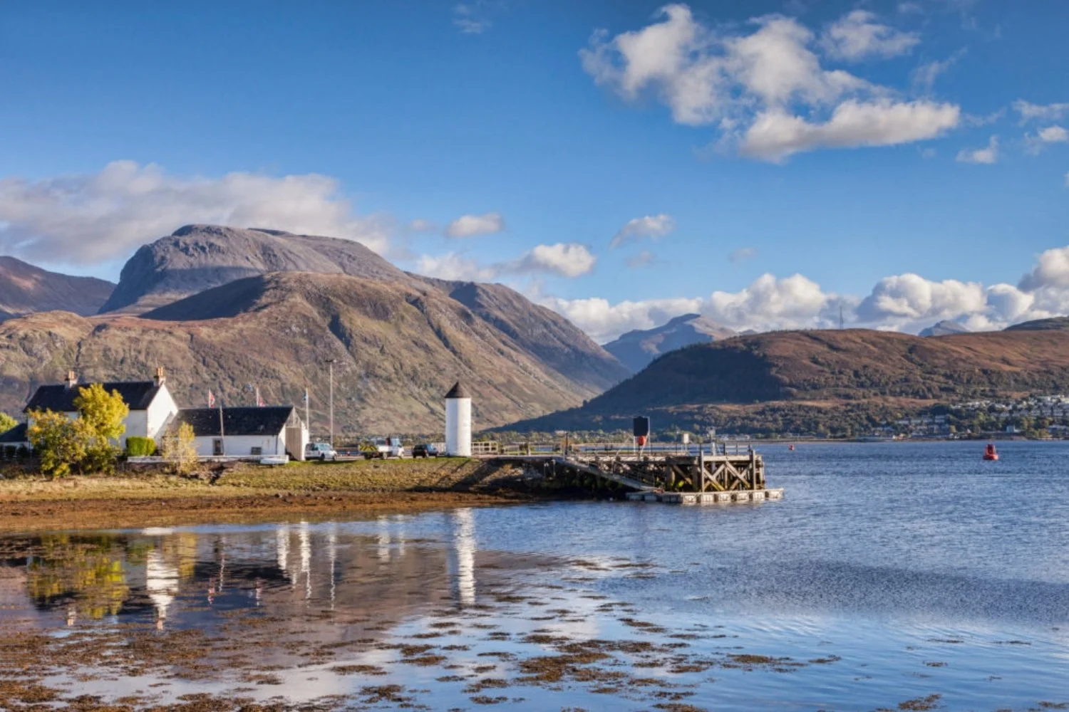A scenic view across the calm waters of Loch Eil showing a white cottage shoreline and pier at Corpach, with the snow-dusted, rugged slopes of Ben Nevis rising majestically into soft sunlight and clouds behind it.