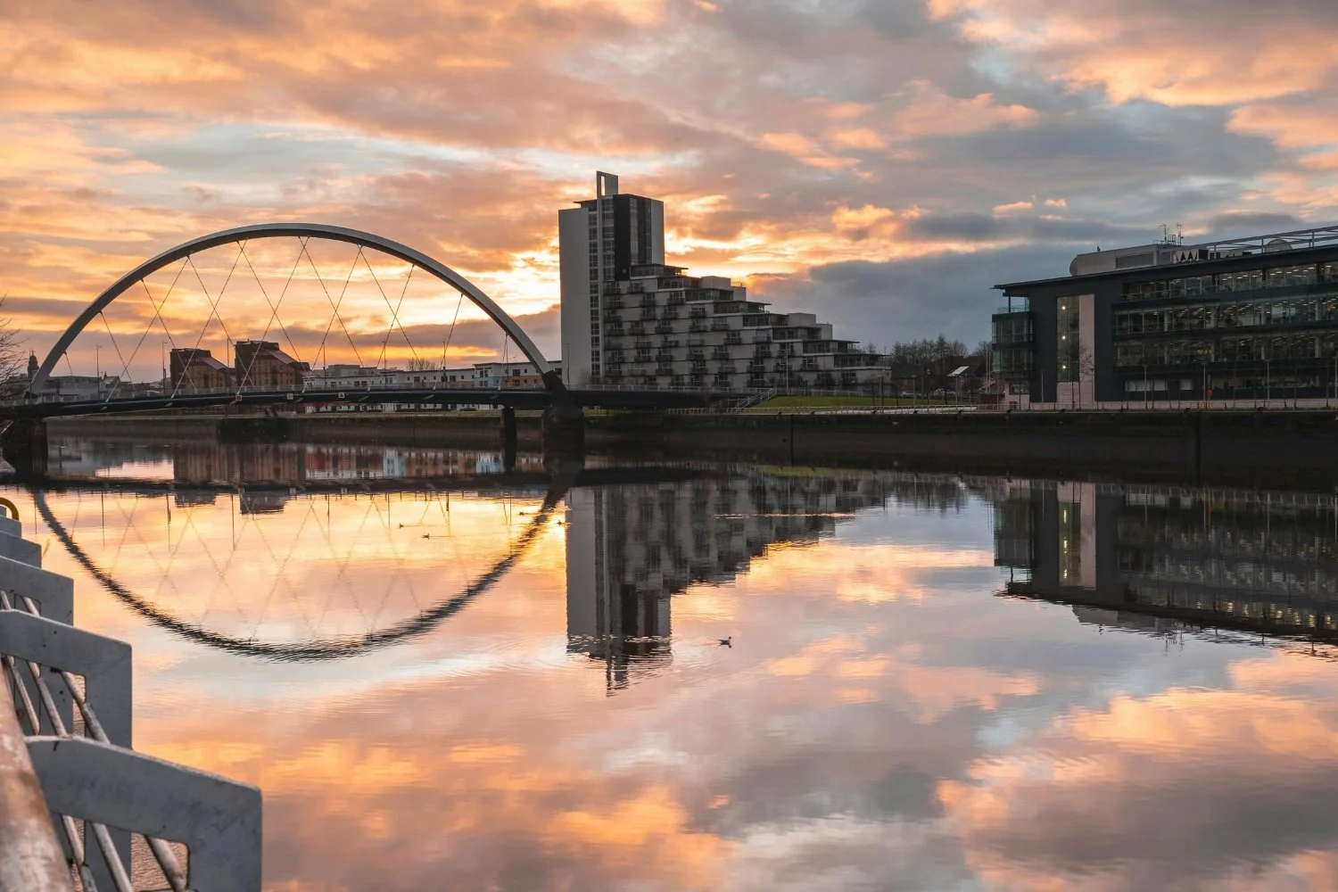 The Clyde Arc bridge in Glasgow beautifully reflected in the glass-like surface of the River Clyde during a warm, golden orange sunset, with modern riverside architecture lining the shore.