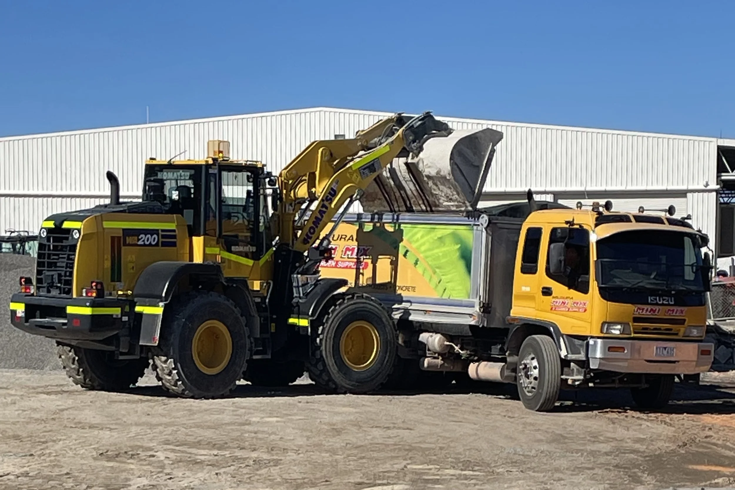 Construction site with a yellow loader lifting gravel into a truck, with a white industrial building in the background.