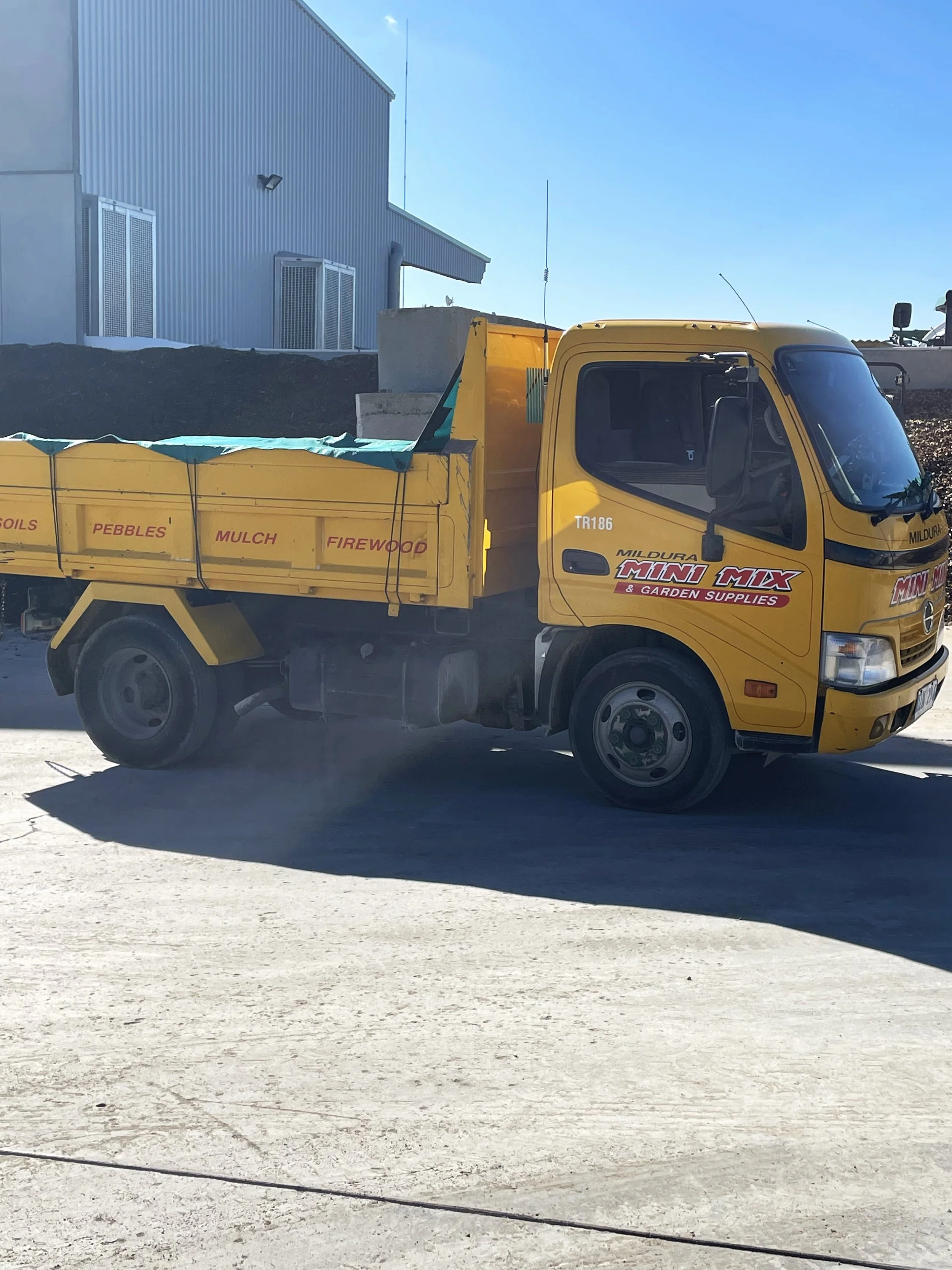 A yellow delivery truck parked outdoors next to a building, with the truck's side showing labels for various building materials like pebbles, mulch, firewood, and soils. The truck is branded with 'Mildura Mini Mix & Garden Supplies.'