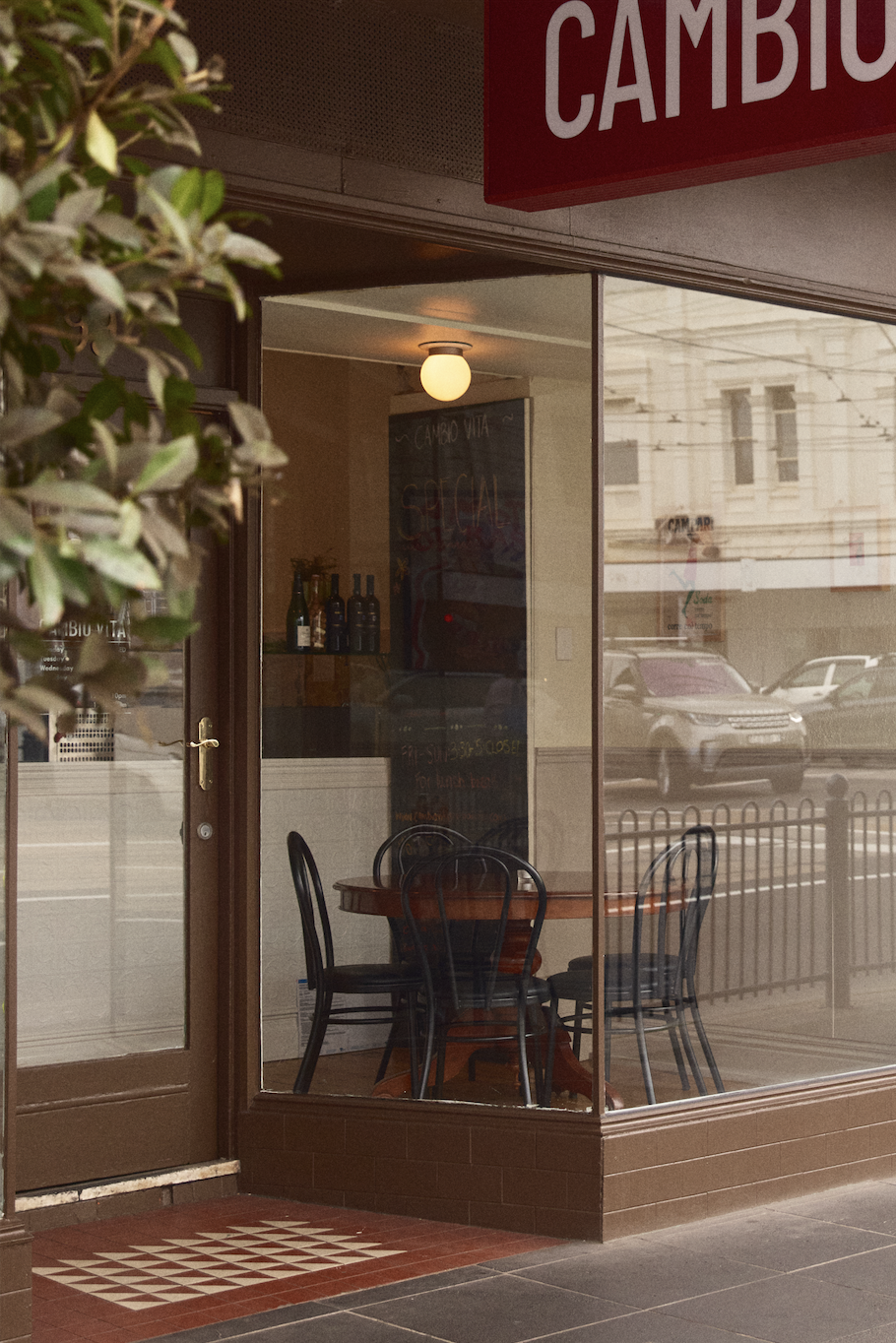 The image shows the exterior and interior of a small restaurant or cafe with a glass front. Inside, there are three black chairs around a wooden table. The outside sidewalk features a decorative tile pattern near the door, and a sign with red and white text partly visible reads "CAMBU". The reflections on the glass show parked cars and surrounding buildings.