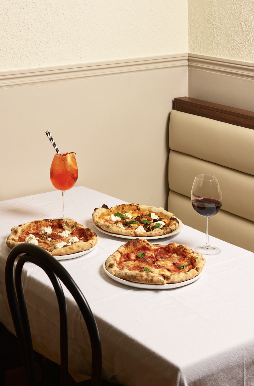 A table set with three pizzas, a glass of red wine, and a cocktail with a straw in a restaurant or dining room.