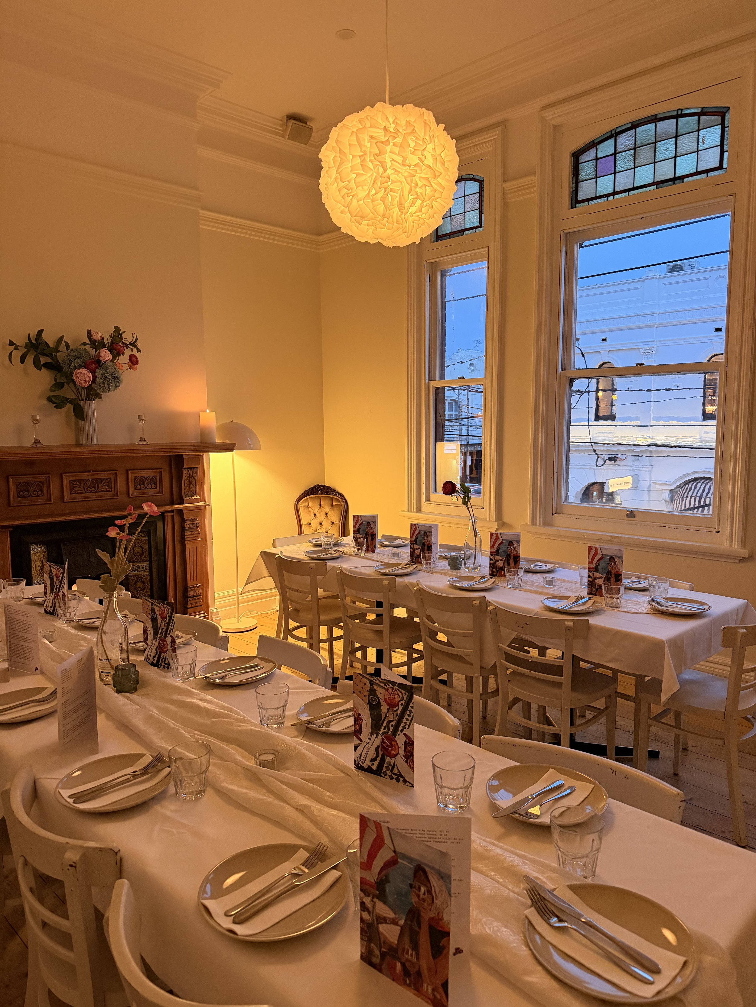 A cozy dining room with a long table set with white plates, silverware, and clear glasses, decorated with a small menu and flower vases. The room has tall windows letting in natural light, a fireplace with flower arrangements on top, and a warm overhead light fixture.