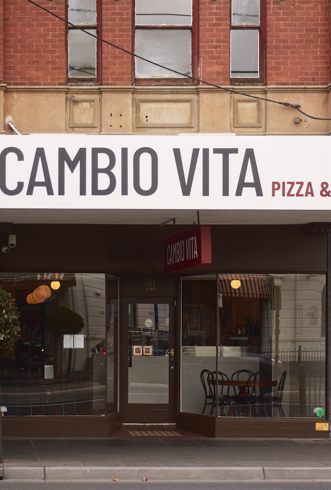 storefront with a white sign reading 'CAMBIO VITA' and 'PIZZA &' in red, large window, entrance door, tables, and chairs inside, above a brick building.