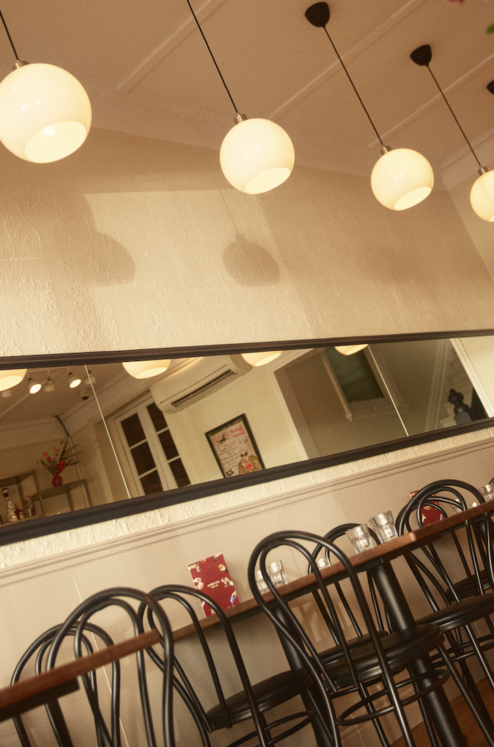 Dining area with a long wooden table, black chairs, drinking glasses, and a large mirror on the wall. Pendant lights hang from the ceiling.
