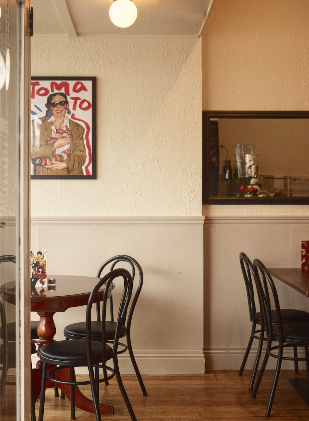 Empty restaurant with a round wooden table, four black chairs, and a framed retro-style artwork of a woman holding a stack of cans on the wall.