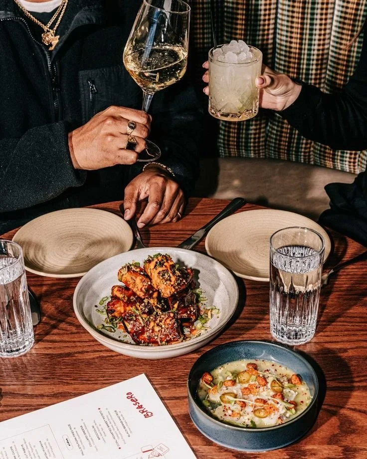 Two people holding glasses of wine and a cocktail at a table set with dishes, water glasses, and a menu.